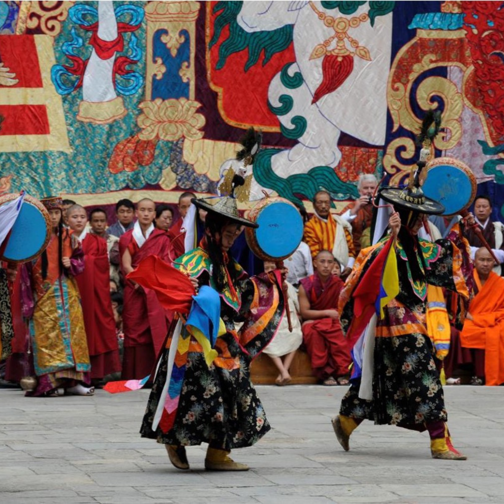 Two individuals in traditional Tibetan attire perform a dance with drums and hats, with a group of monks and onlookers in robes in the background against a large, colorful thangka tapestry.