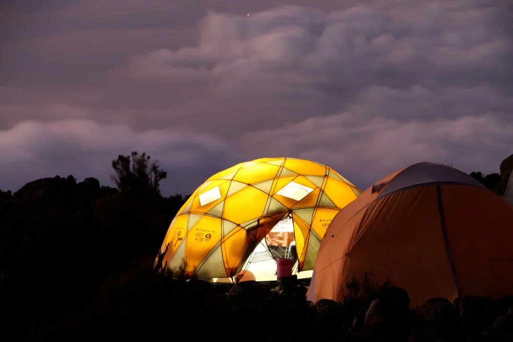 Two tents set up outdoors during twilight, one illuminated from inside, against a cloudy sky and silhouette of trees.