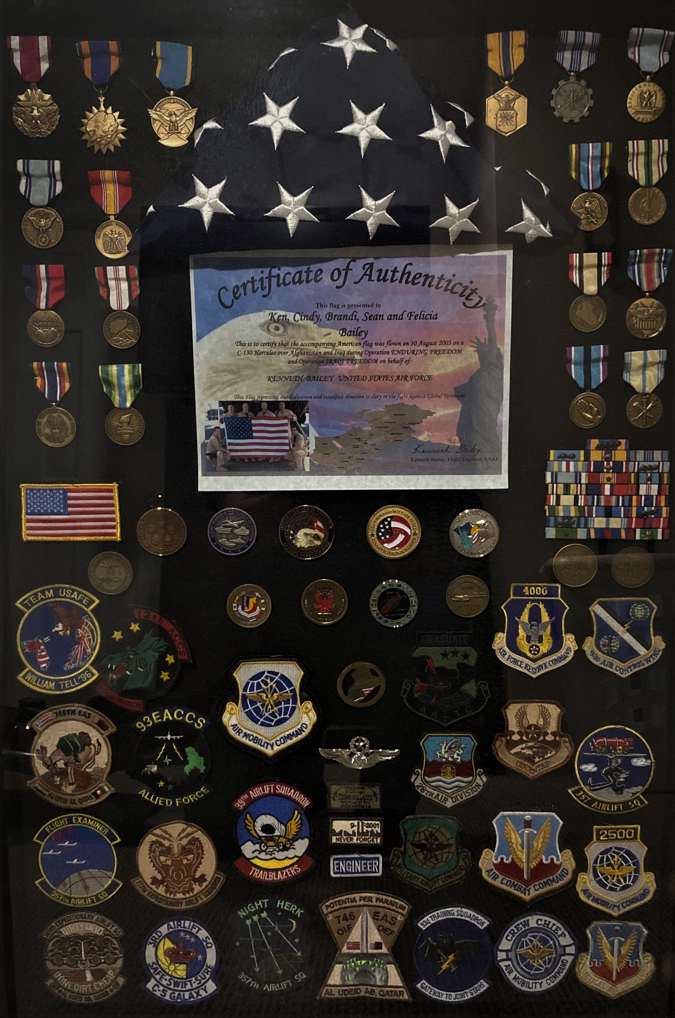 Display case with numerous military medals, patches, and a certificate of authenticity. The case features a fabric with white stars on a dark background, resembling the American flag.