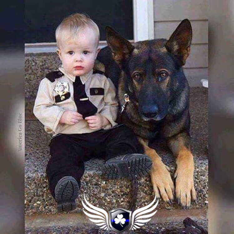 A young boy dressed as a police officer sitting on the ground next to a large black and brown dog, both looking at the camera, outside a house.