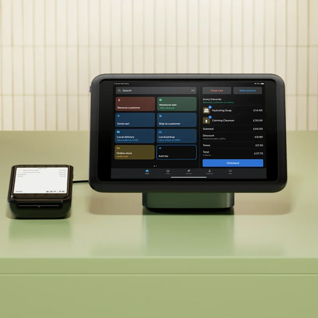 A digital tablet displaying an online shopping checkout screen and a small card reader on a light green table against a tiled wall.