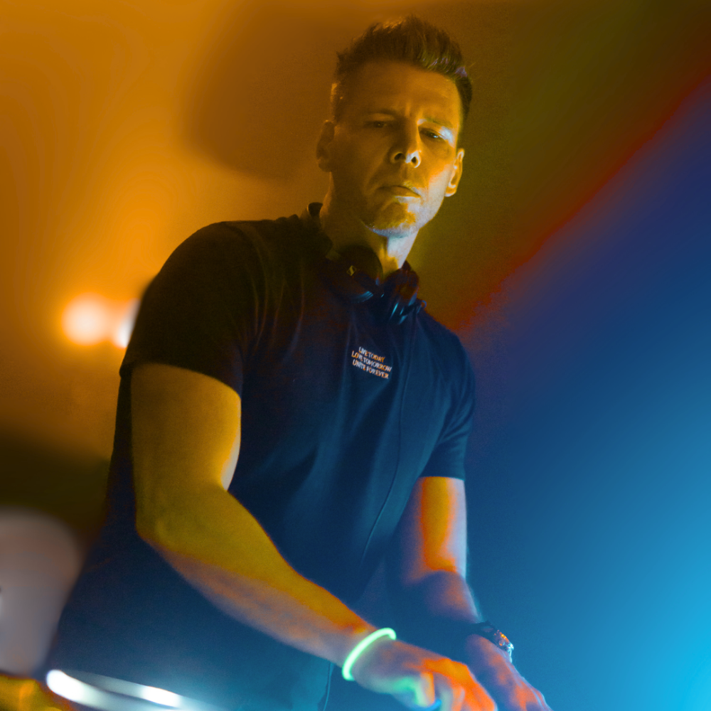 A DJ performing with colorful lighting effects, wearing a black T-shirt and headphones around his neck.