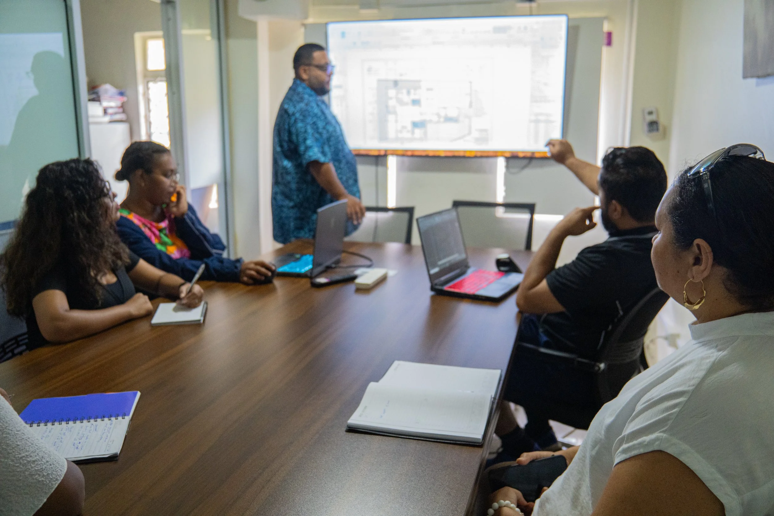 A diverse group of five people in a meeting room attending a presentation. One person is standing next to a large screen displaying a diagram or chart, while the others are seated around a wooden table with laptops, notebooks, and smartphones.