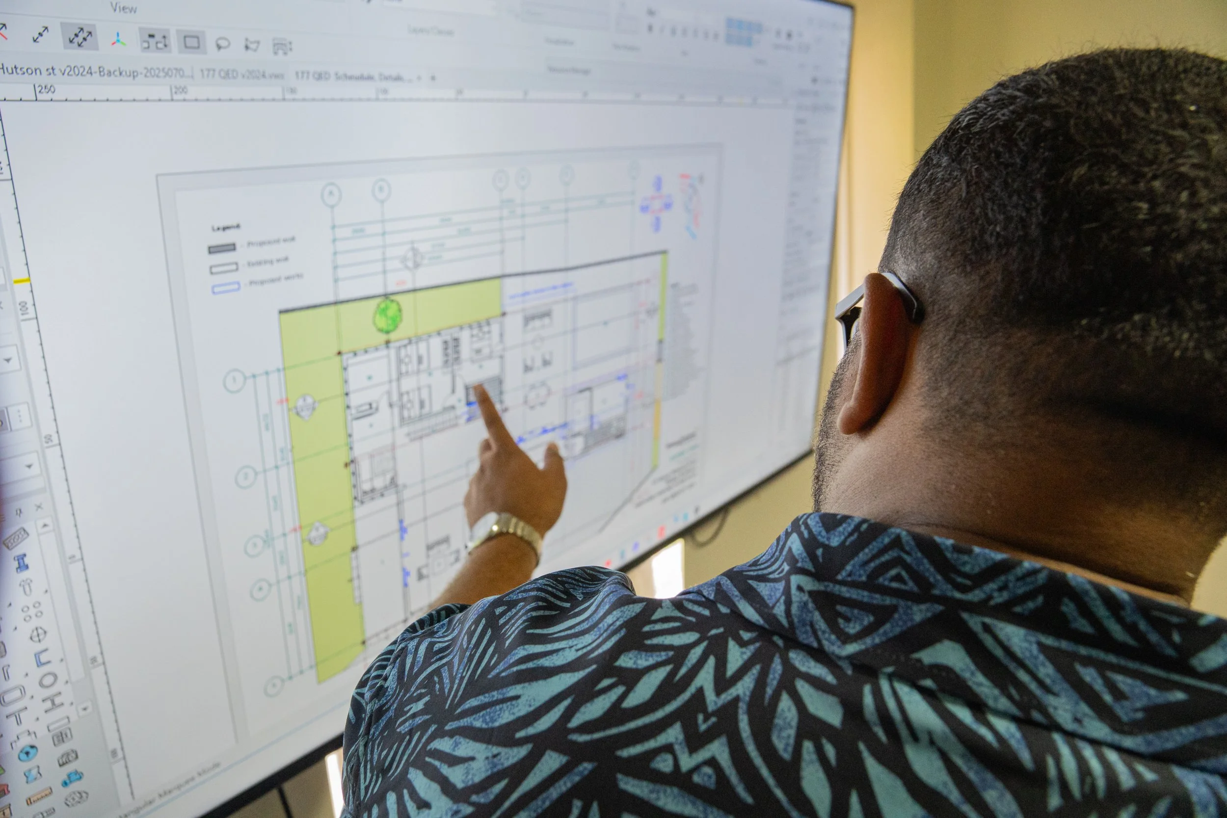 A man with glasses and a patterned shirt pointing at a computer screen displaying a technical drawing or architectural plan.