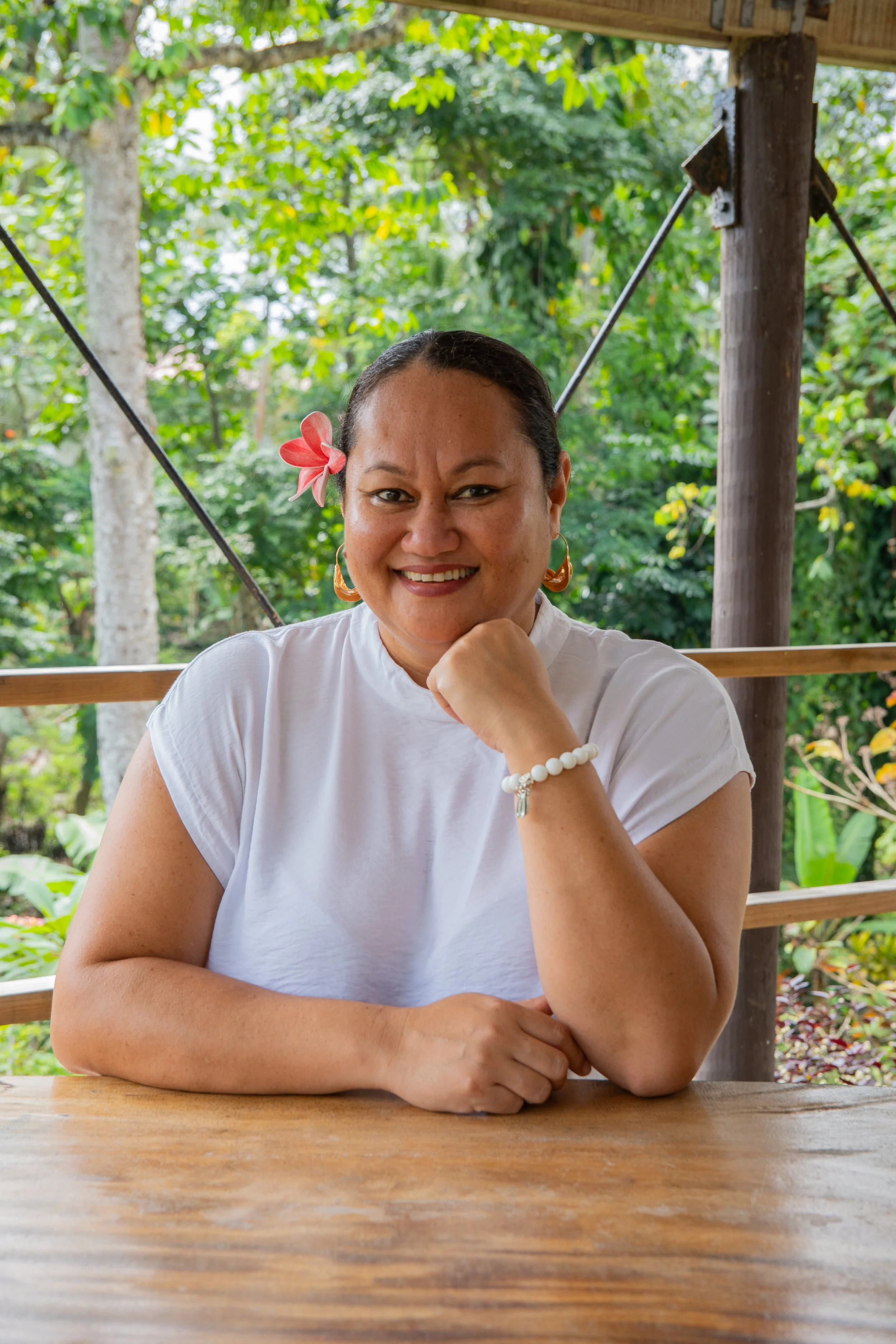 Smiling woman with dark hair, wearing a white t-shirt, accessorized with a pink flower in her hair, earrings, and bracelet, sitting outdoors at a wooden table in a lush green setting.