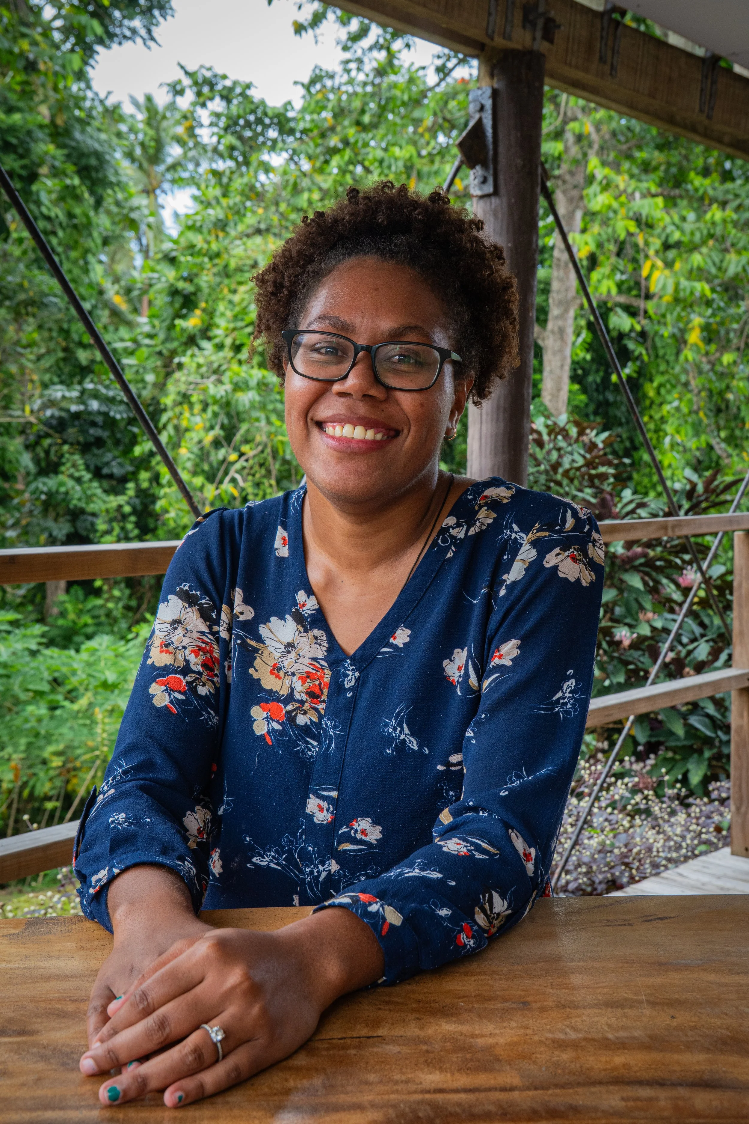 A woman sitting at a wooden table outdoors, smiling, with glasses, wearing a navy blue blouse with floral patterns, and surrounded by green trees.