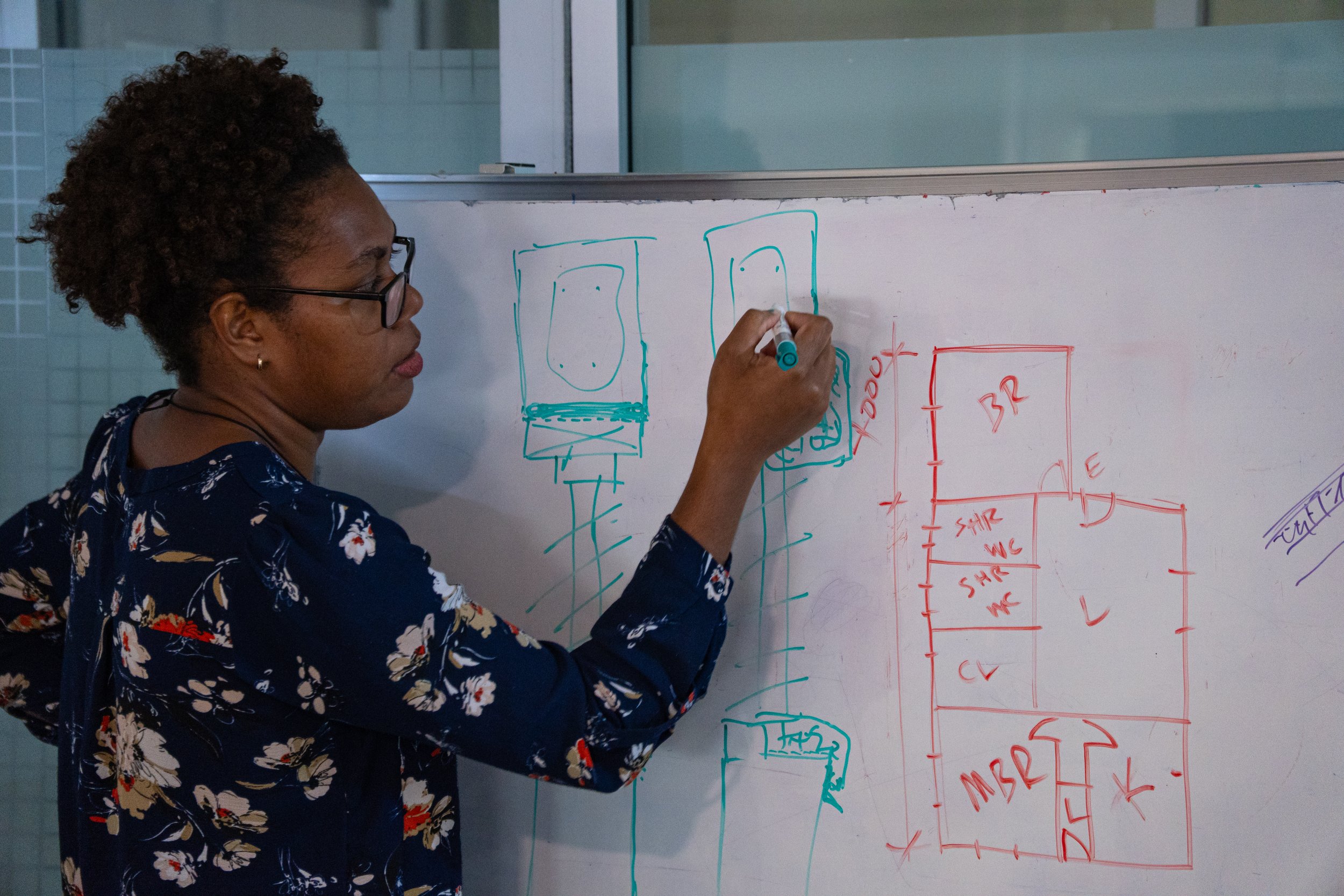 A woman wearing glasses and a floral shirt is writing on a whiteboard with a blue marker. The whiteboard has technical sketches and floor plans drawn in blue and red ink.