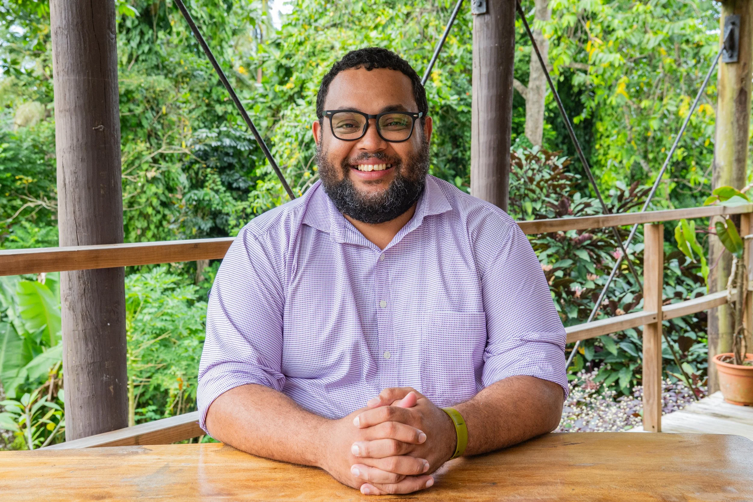 A smiling man with glasses and a beard sitting at a wooden table outdoors surrounded by greenery.