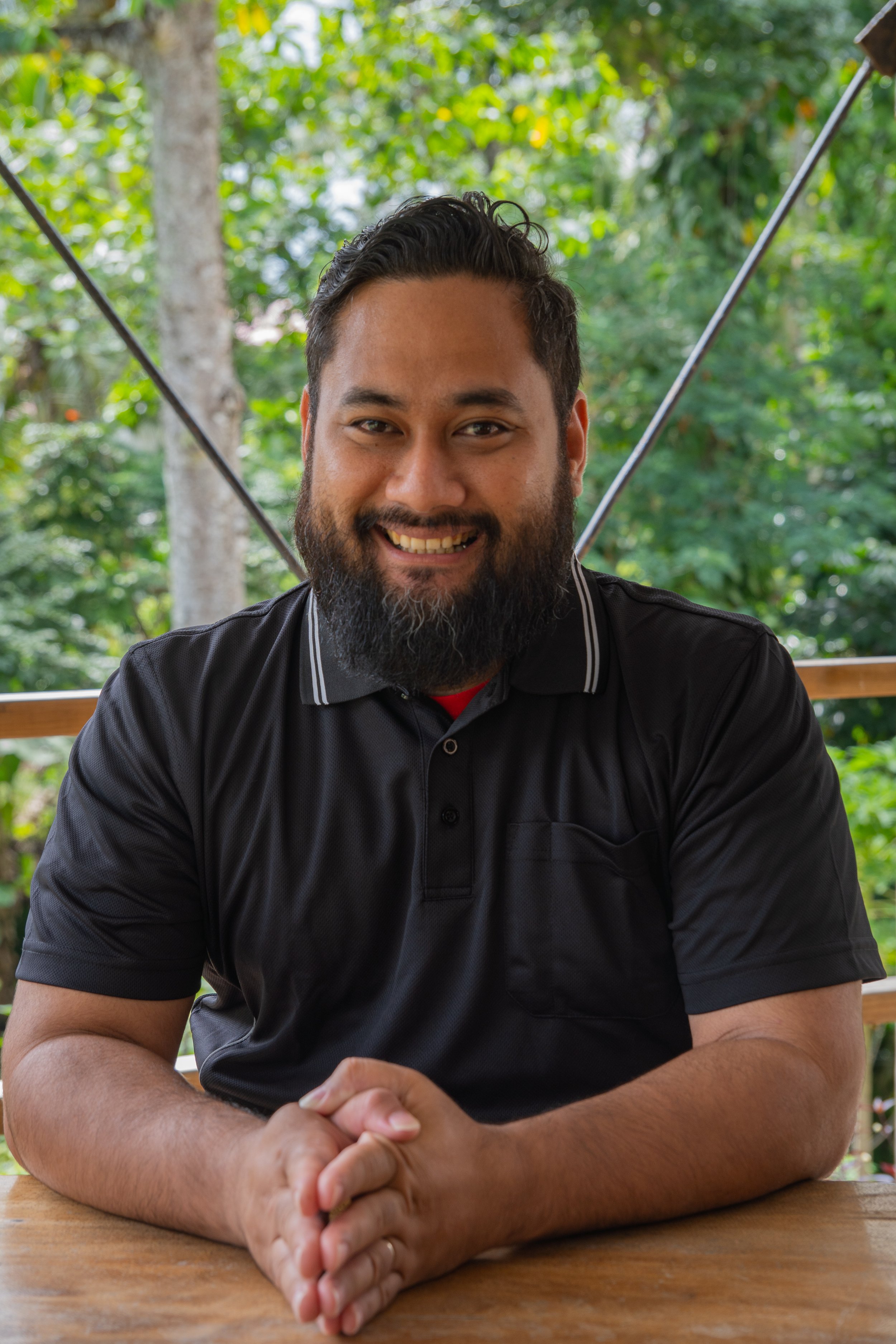 A man with a beard and dark hair, wearing a black polo shirt, smiling and sitting at a wooden table outdoors with green foliage in the background.