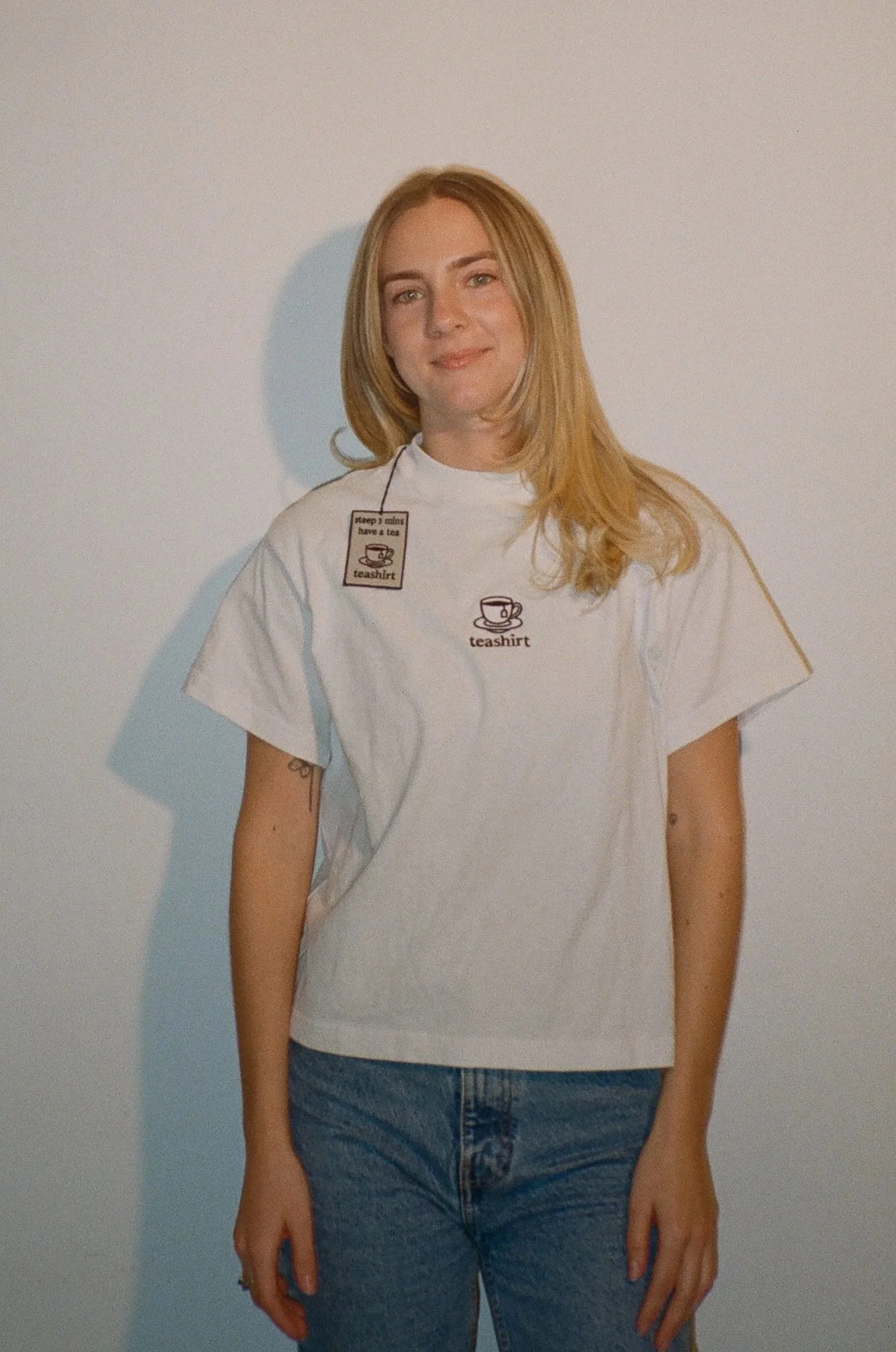 A young woman with blonde hair wearing a white t-shirt with a tea cup graphic and the phrase "teashirt, sleep's dreams have a tea" against a plain white background.
