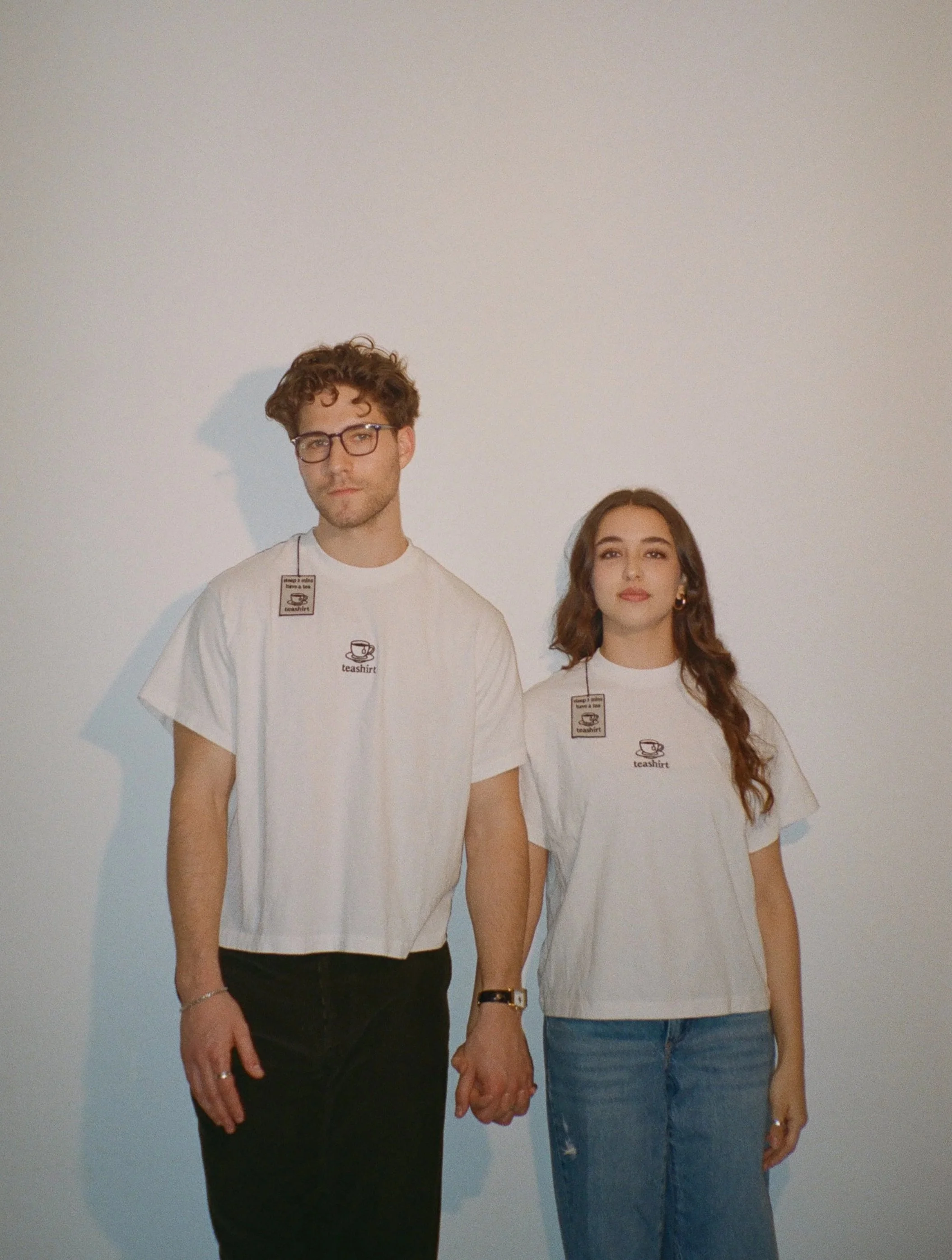 A young man and woman holding hands, both wearing white t-shirts with a coffee cup logo and the word "teashirt," standing against a plain white wall.