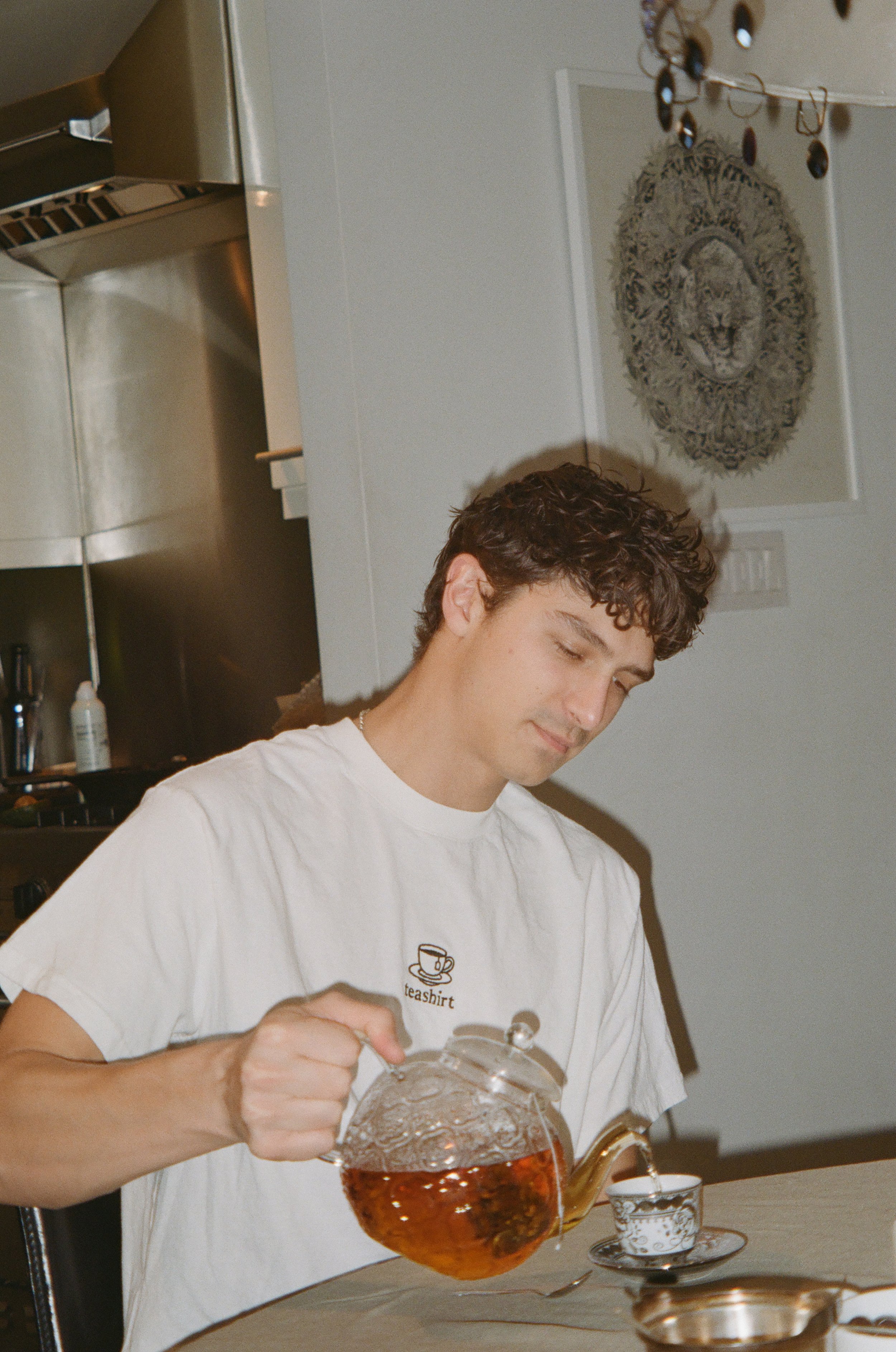 A young man with curly brown hair pouring tea from a glass teapot into a teacup on a table.