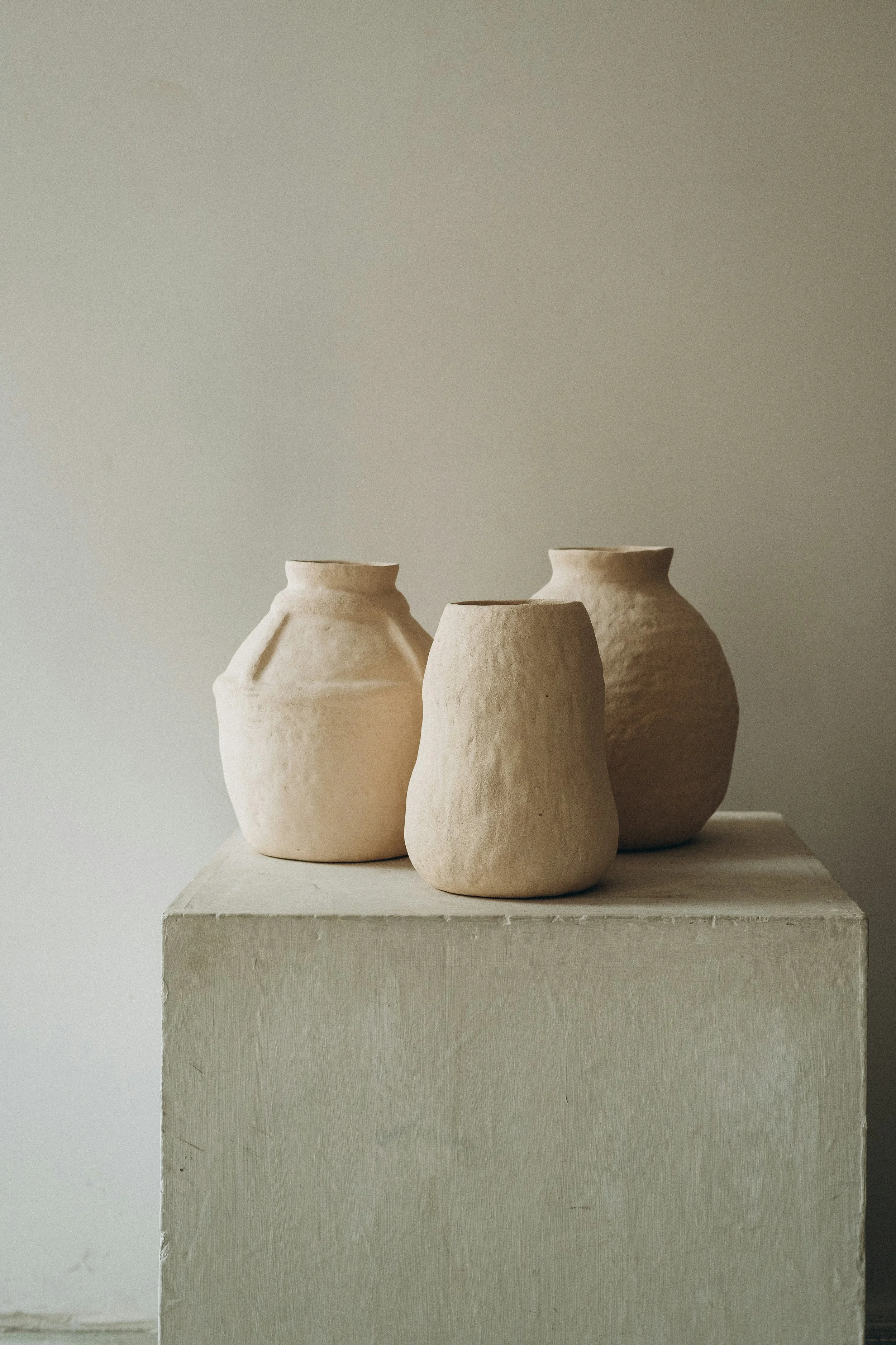 Three cream-colored ceramic vases of varying shapes on a white pedestal against a plain wall.