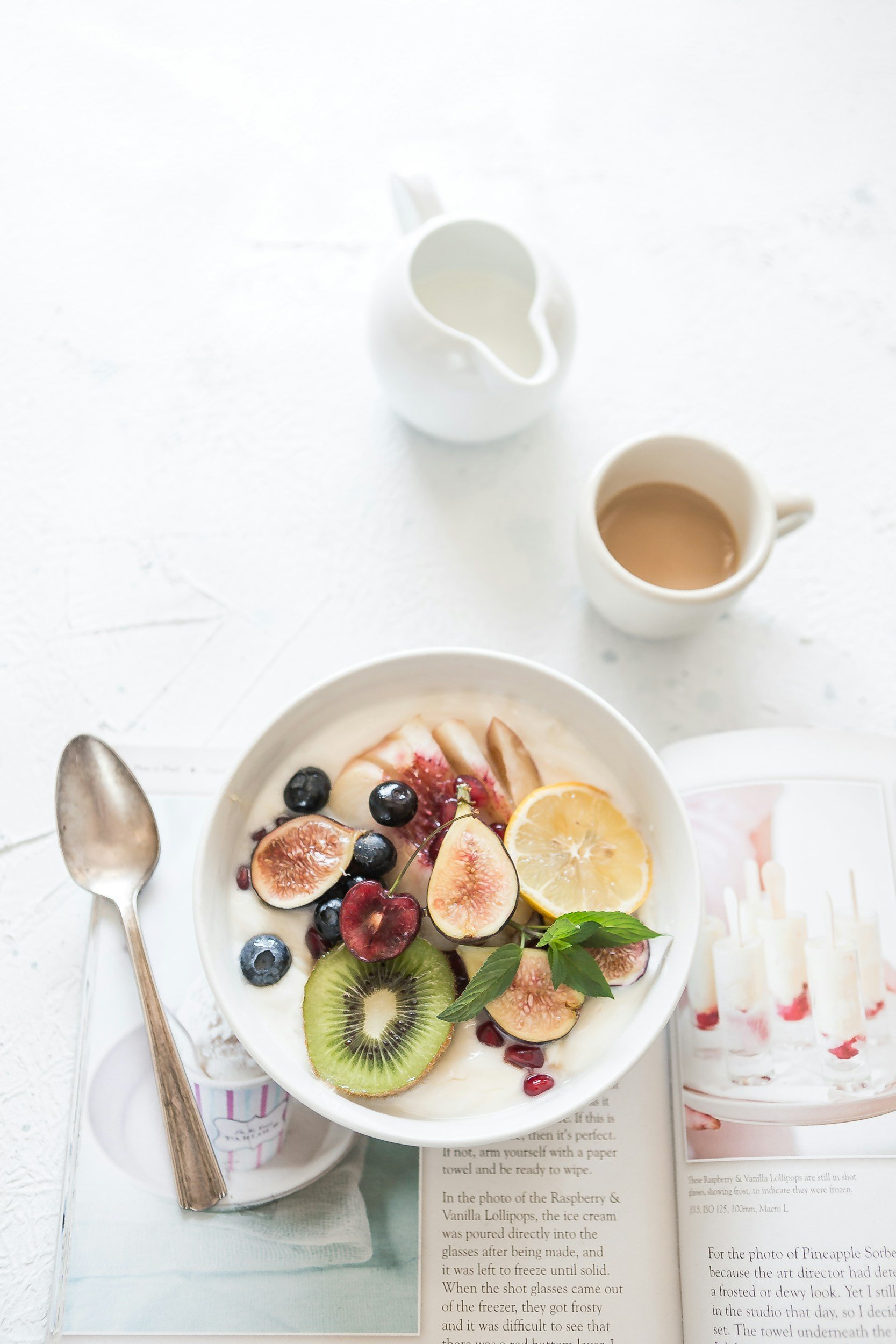 A bowl of yogurt topped with fresh fruits including kiwi, blueberries, figs, cherries, and a lemon slice, with mint leaves. Two small white pitchers, one empty and one with a brown liquid, are placed above the bowl. An open book and a silver spoon are also visible.