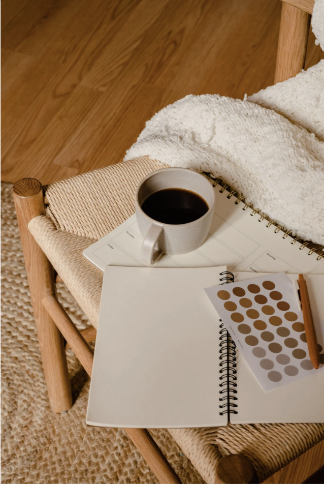 Coffee cup, open notebook, color swatches, and pen on a small wooden chair with a textured blanket, next to a rug and wooden flooring.