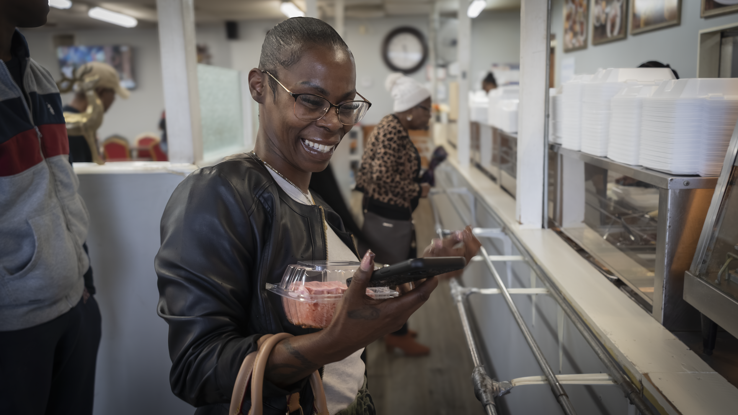 A woman with glasses and a leather jacket is smiling and looking at her phone while holding a clear plastic container of raw meat in a grocery store. Other people are visible shopping in the background.