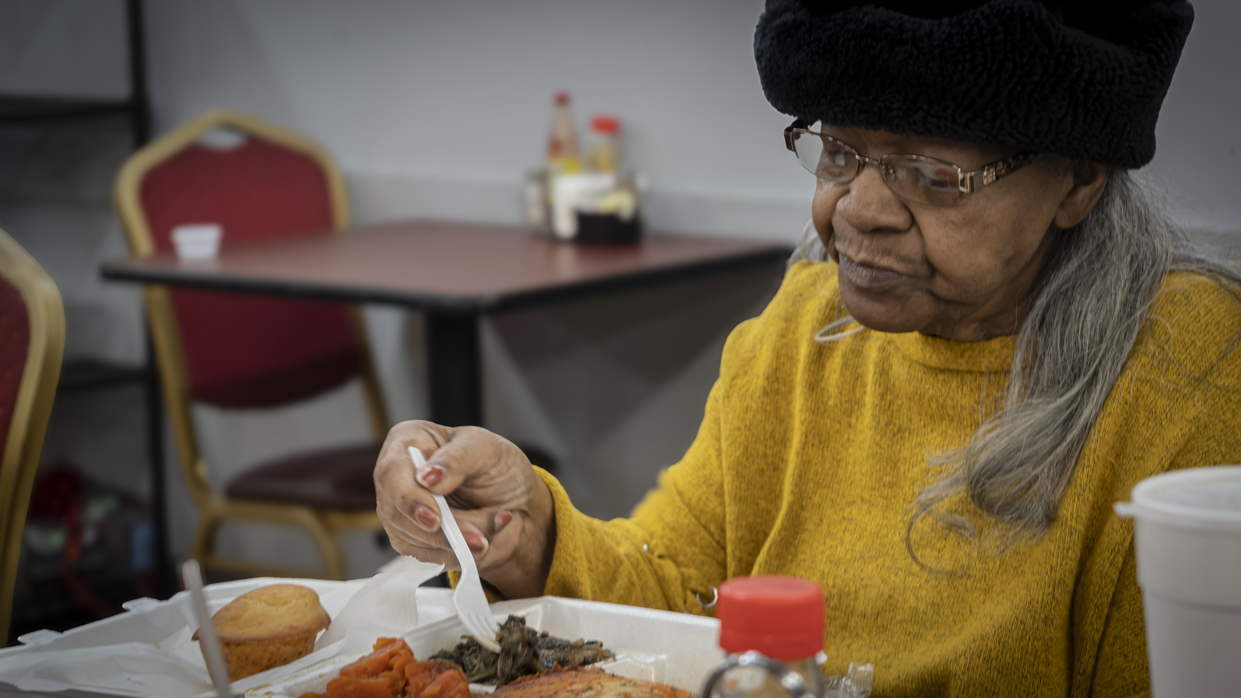 An elderly woman in glasses and a black hat, wearing a yellow sweater, is eating food from a white takeout container at a dining table.