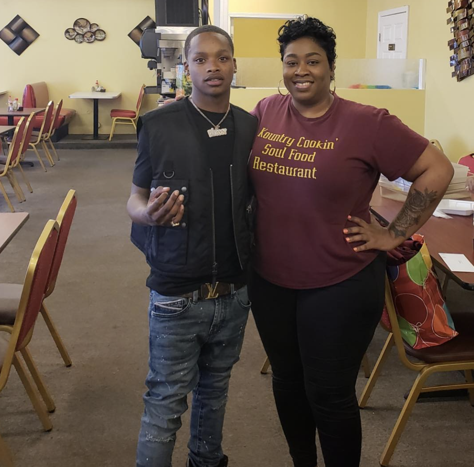 A young man with a short haircut wearing a black vest, gray jeans, and multiple necklaces poses with a woman with short curly hair, wearing a maroon T-shirt with the words 'Kountry Cookin' Soul Food Restaurant' printed on it. They are standing inside