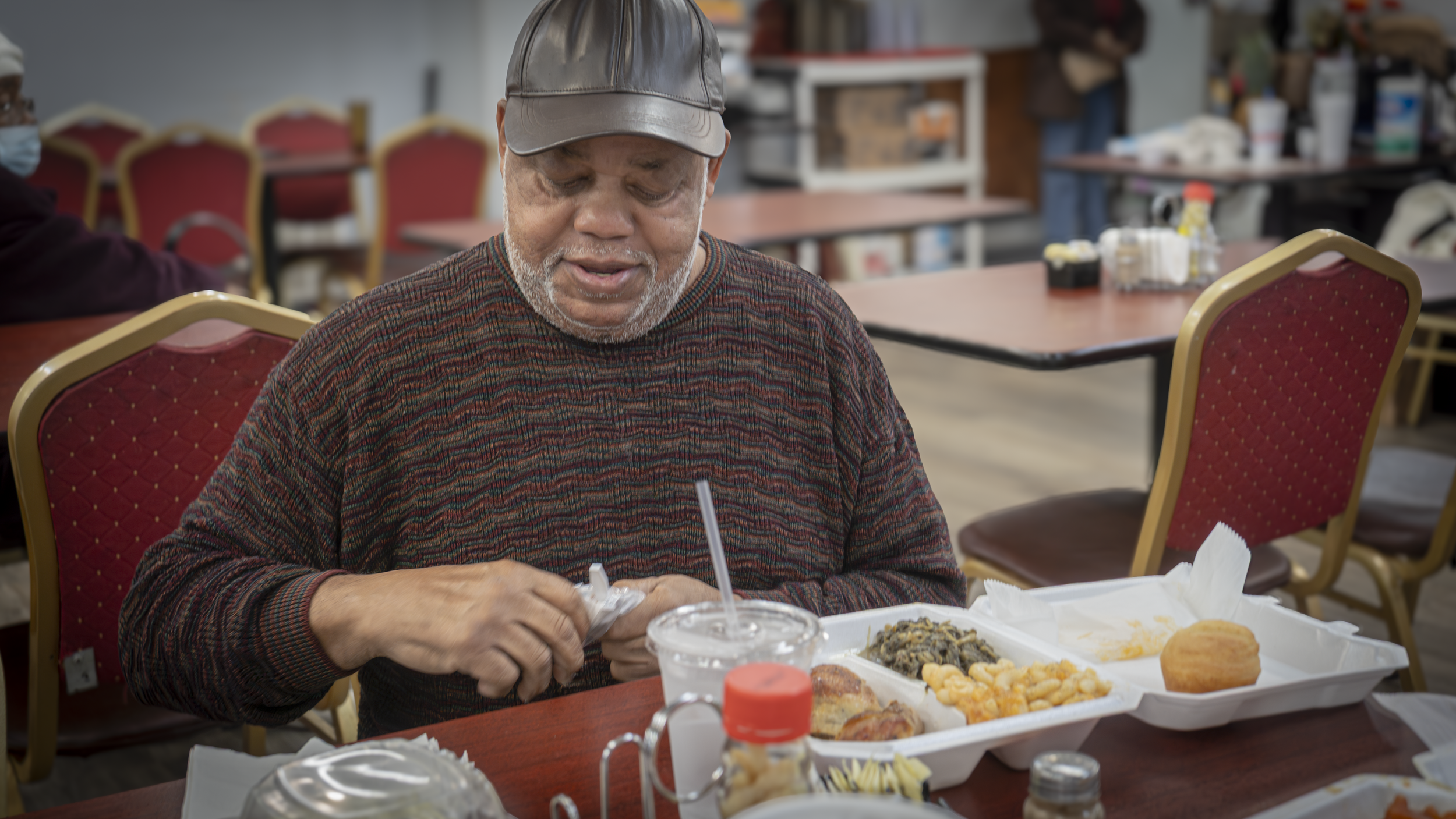 An elderly man with gray hair and a beard, wearing a cap and colorful sweater, is sitting at a table with a styrofoam takeout container of food, a drink, and condiments.