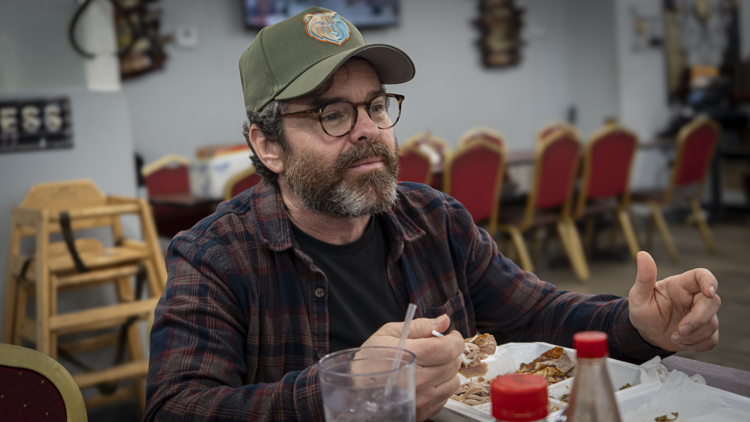 A man with glasses and a beard, wearing a green cap with a logo, sitting at a table in a restaurant or cafeteria, eating food from a white tray.