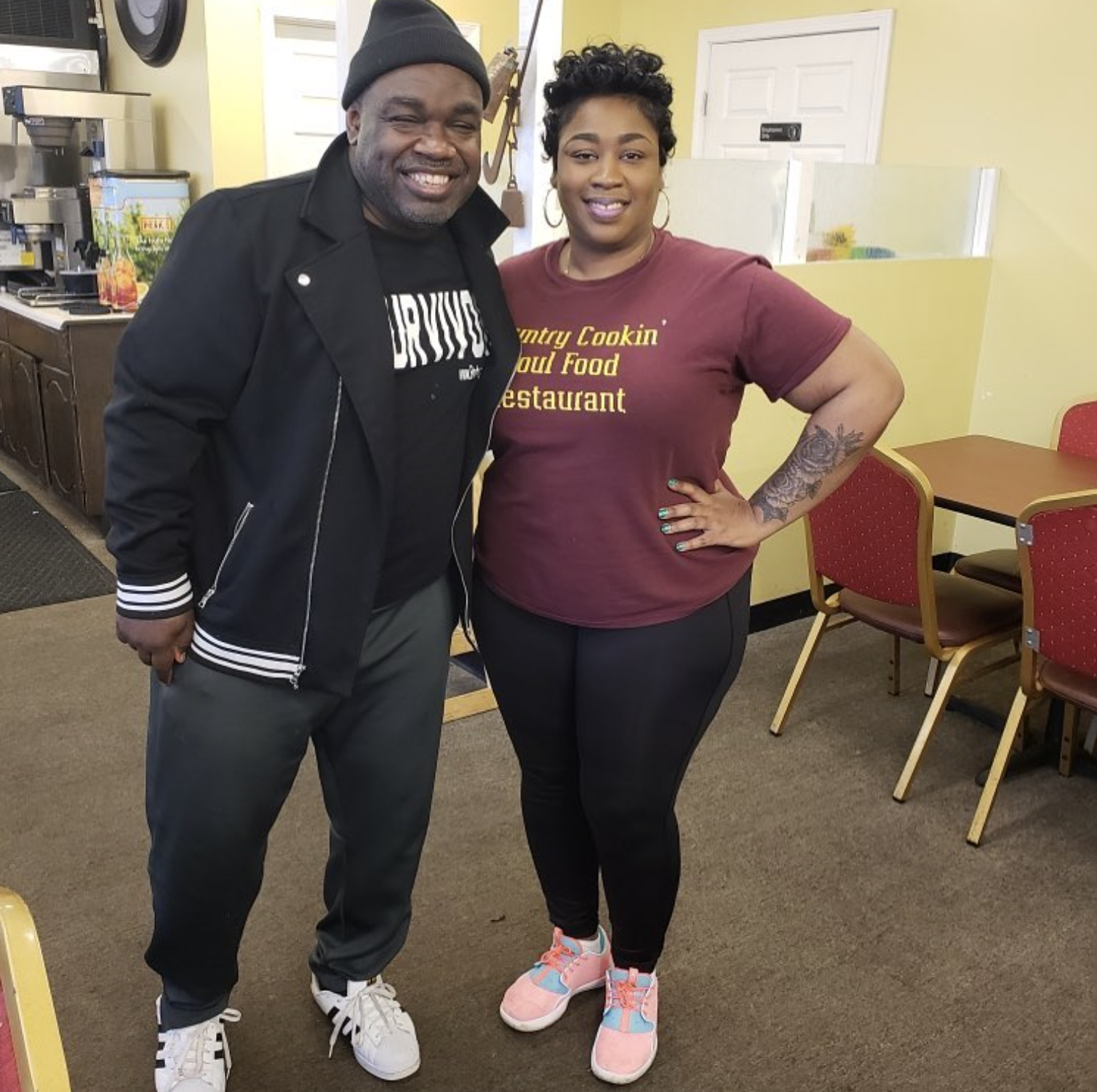A man and woman standing together inside a restaurant, smiling at the camera. The man is wearing a black jacket, gray pants, white sneakers, and a black beanie. The woman is wearing a maroon T-shirt with yellow text, black leggings, and pink sneakers