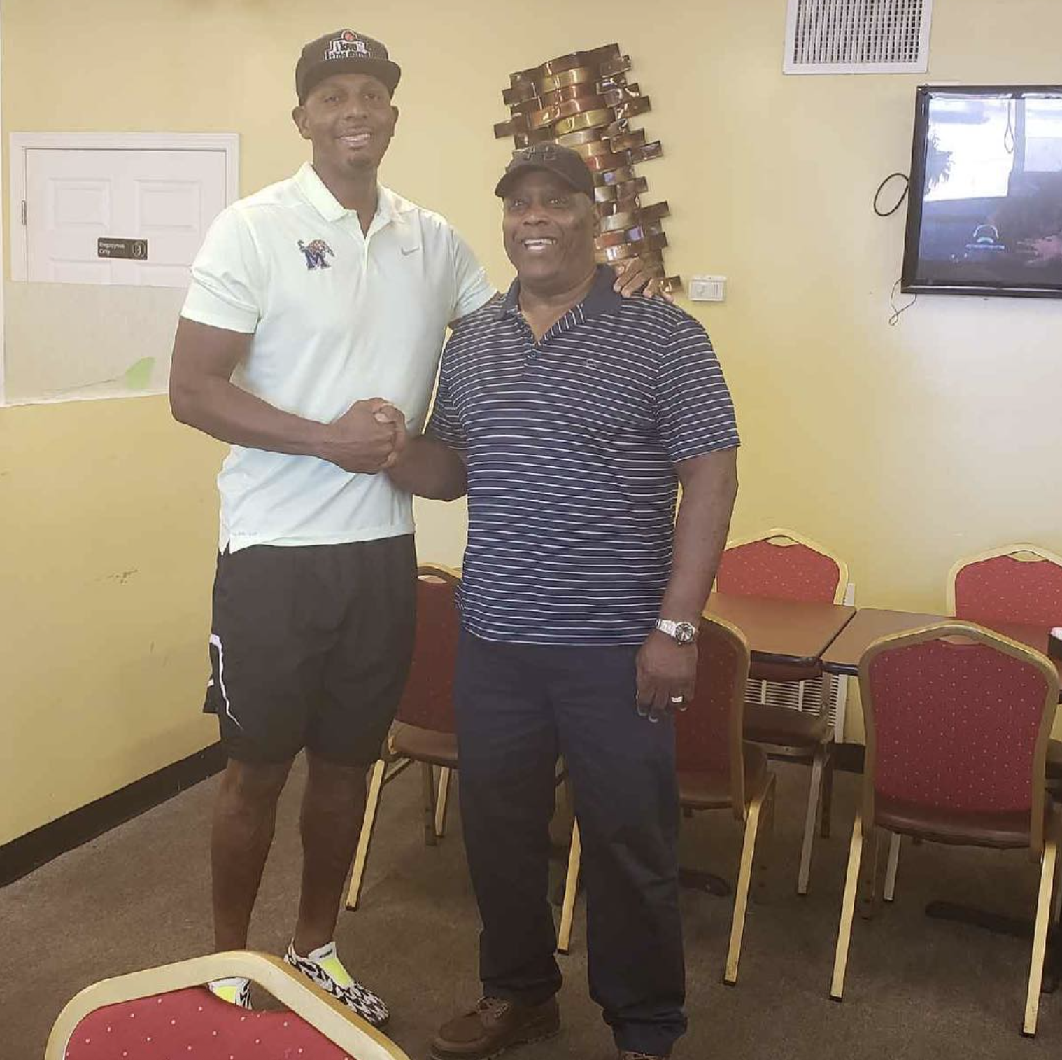 Two men standing together in a room, shaking hands and smiling. One is taller and wearing a white polo shirt, the other shorter wearing a striped polo shirt. There are chairs and a table in the background.