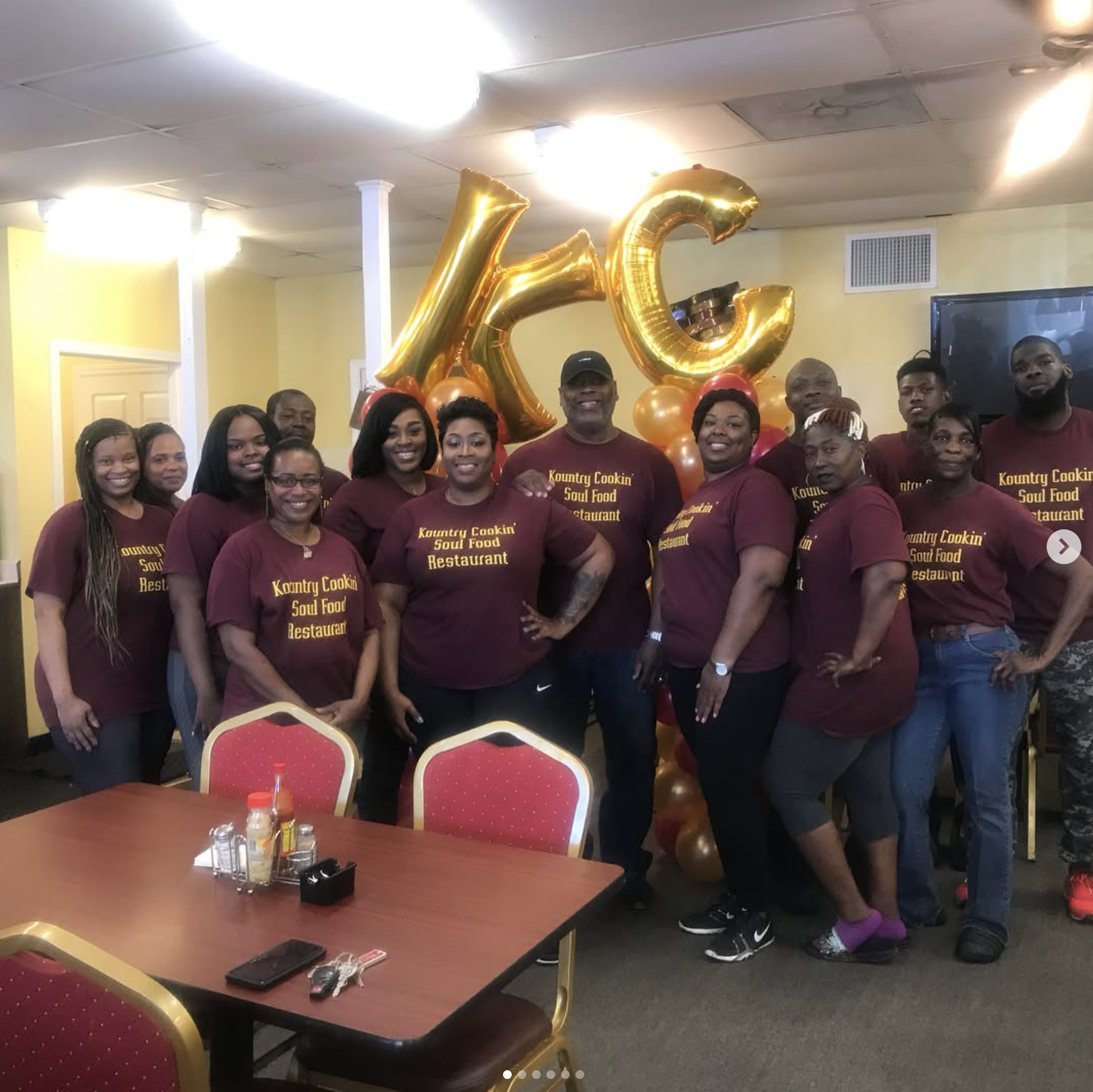 Group of people wearing maroon shirts with yellow text standing in a restaurant, celebrating with large gold balloons spelling 'KC' in the background.