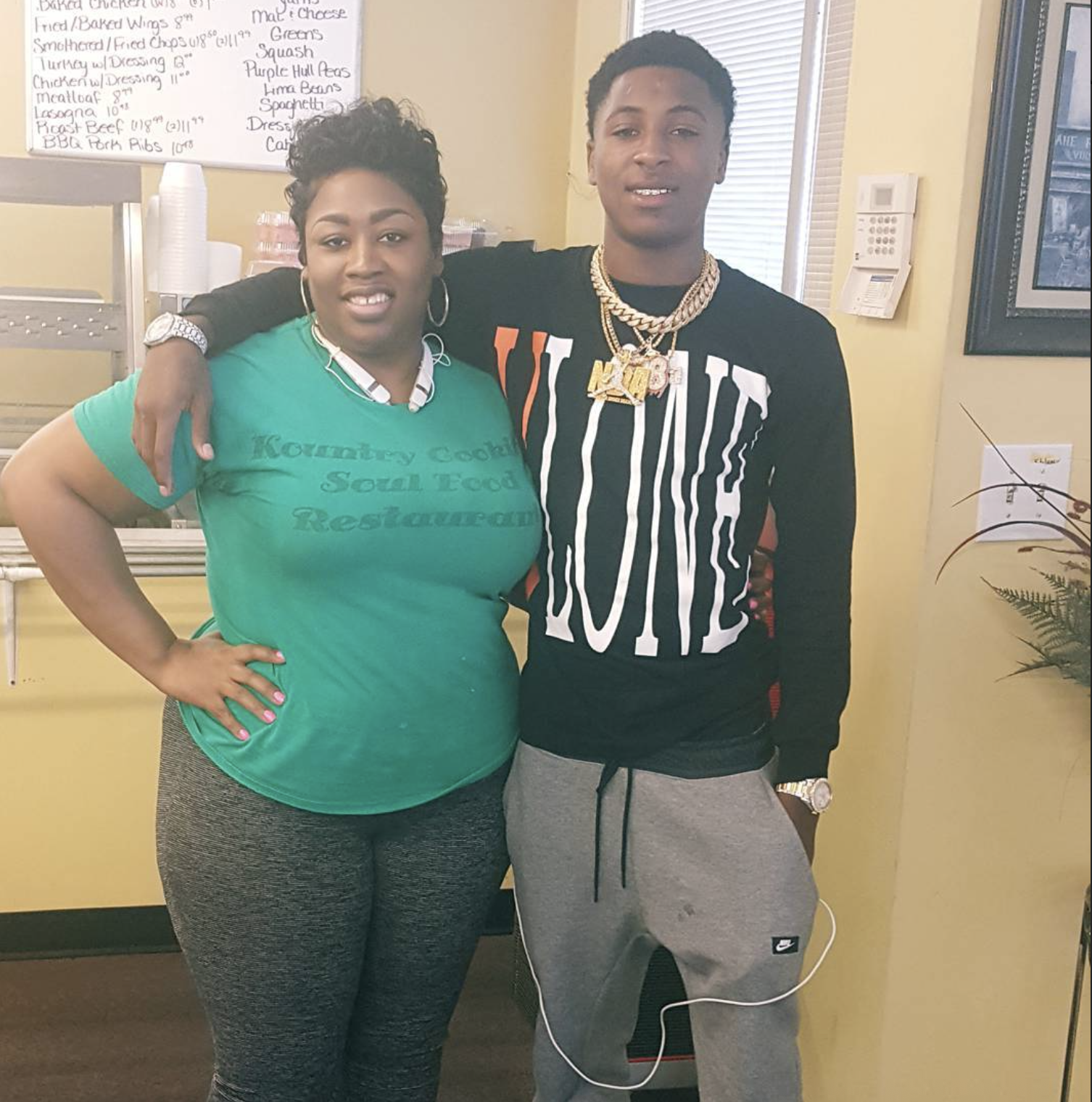 A woman and a young man standing indoors, close together, with the woman having her arm around the young man's shoulders. The woman is wearing a green t-shirt that reads 'Kountry Cook SouL Food Restaurant,' grey patterned leggings, a necklace, and ea