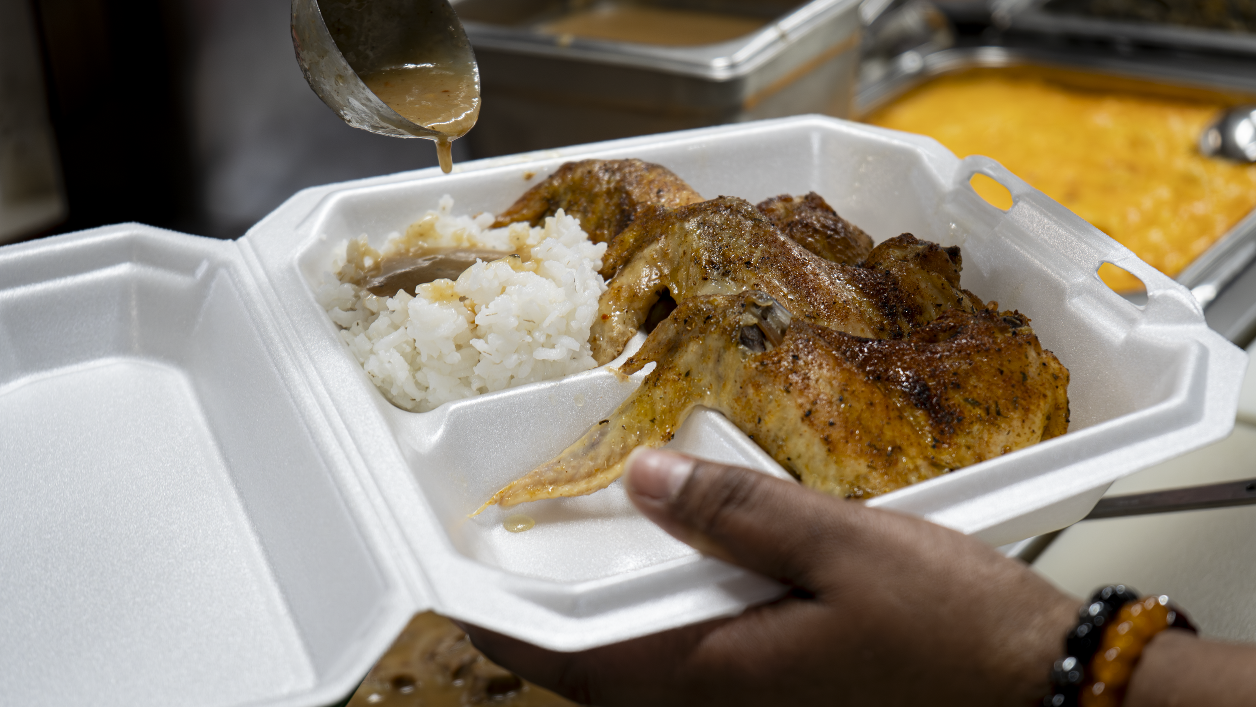 A person holds a foam takeout container containing cooked chicken drumsticks, mashed potatoes with gravy, and a serving of rice. A spoon is pouring gravy onto the rice and chicken.