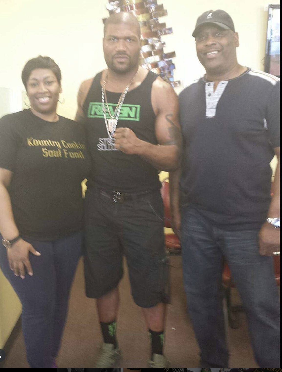 Three people standing together indoors: a woman on the left wearing a black T-shirt with gold lettering, a man in the middle wearing a sleeveless black shirt with green lettering and showing tattoos and a necklace, and another man on the right wearin