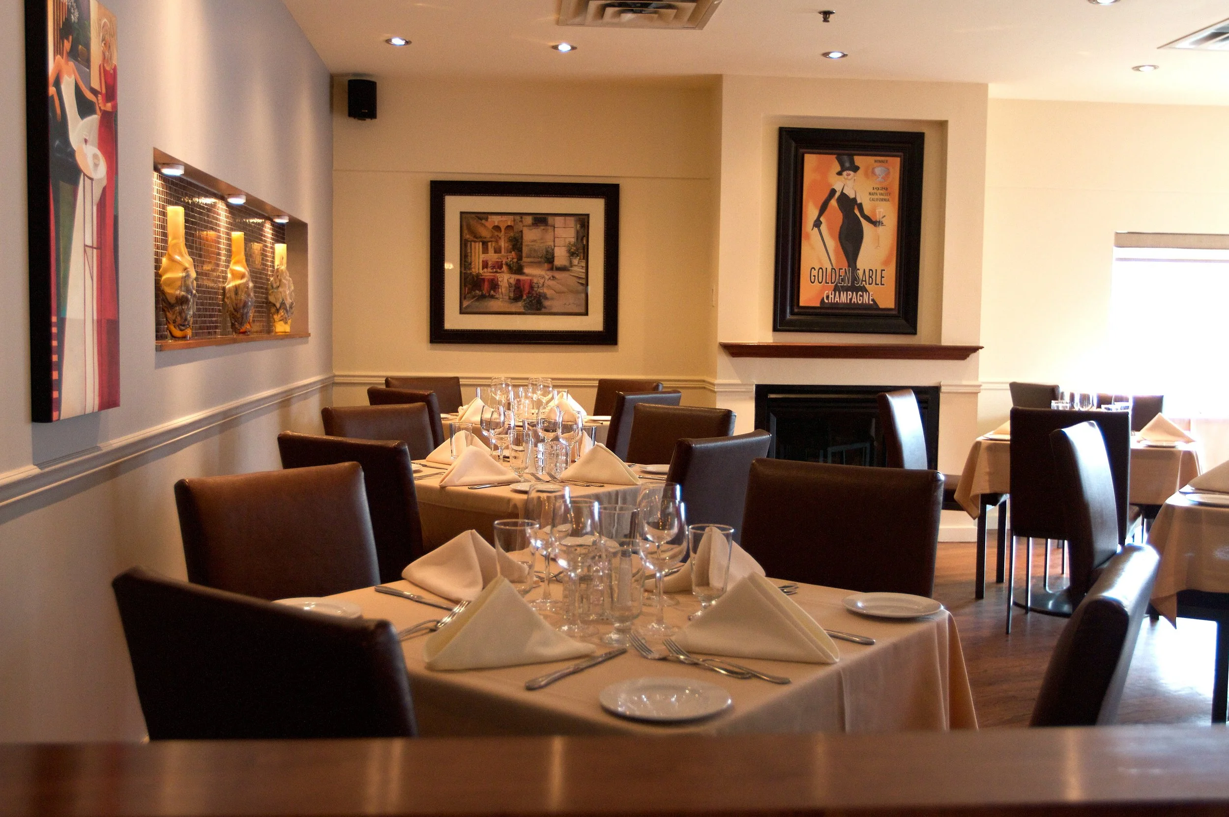 A restaurant dining area with neatly set tables, white tablecloths, glasses, napkins, and silverware. The walls feature framed artwork and decorative displays, and there is a fireplace under one of the frames. Sunlight filters through a window on the right.