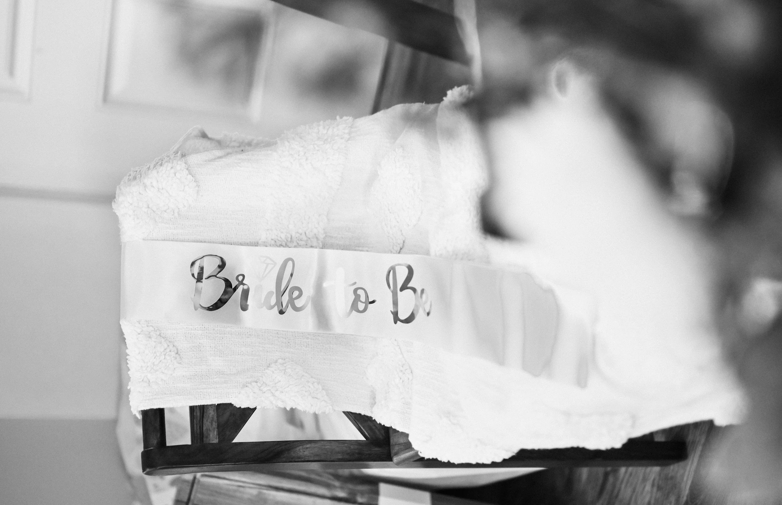 A wooden chair with a torn paper banner that reads 'Bride to Be' in cursive, placed on a table beside a textured white cloth.