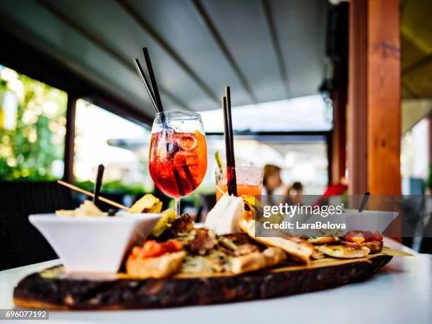 Two colorful cocktails with straws on a wooden serving tray, along with a bowl of chips in the background, set on a table under a covered outdoor area at a restaurant.