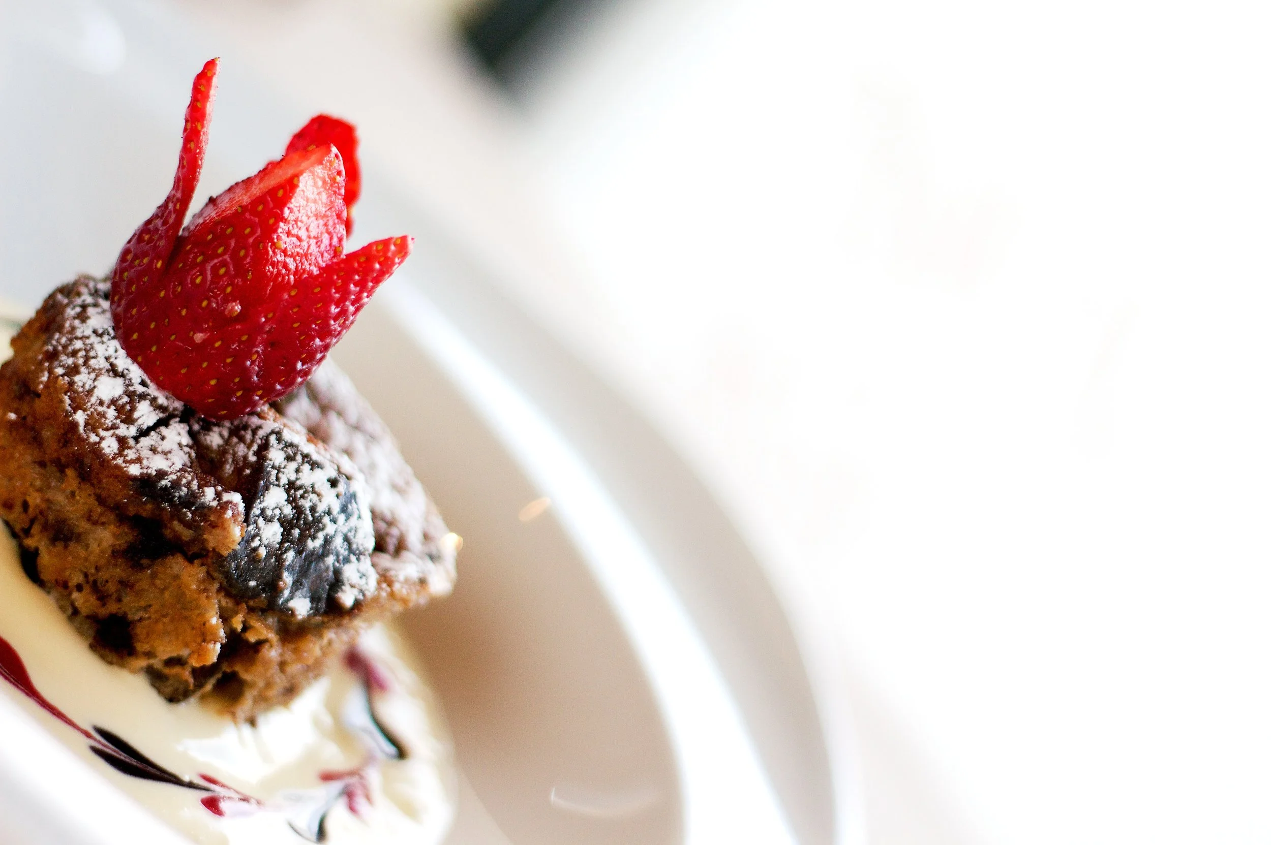 Close-up of a chocolate brownie topped with powdered sugar and a sliced strawberry on a white plate with a decorative red and black sauce