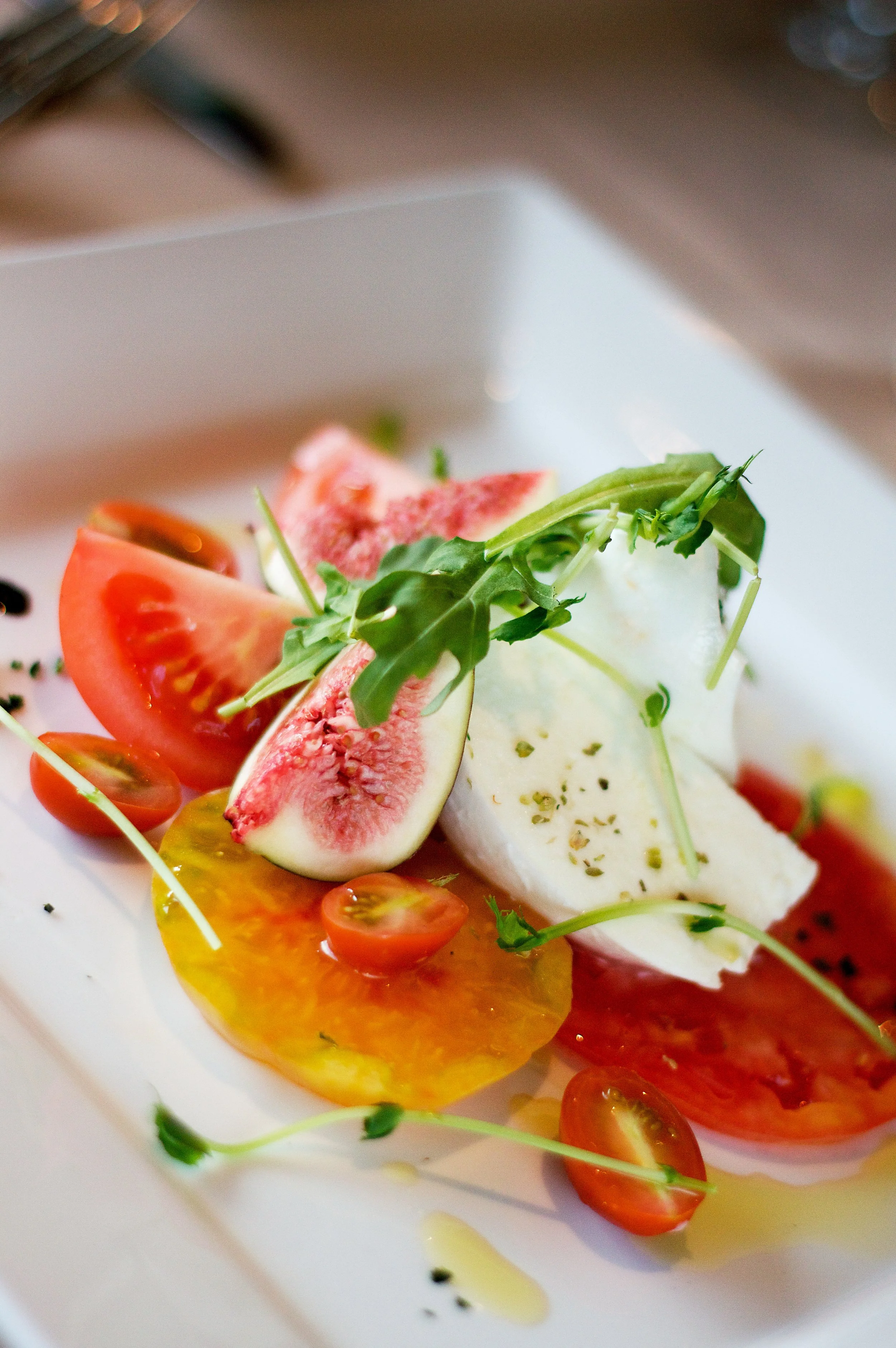 Fresh tomato slices, a fig, cheese, and microgreens on a white plate.