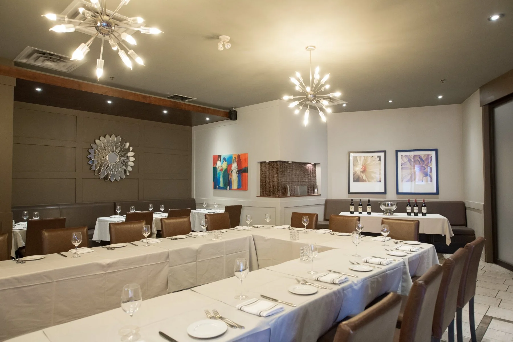 Empty restaurant dining area with white tablecloths, glassware, and cutlery, decorated with paintings on the walls and modern chandeliers hanging from the ceiling.