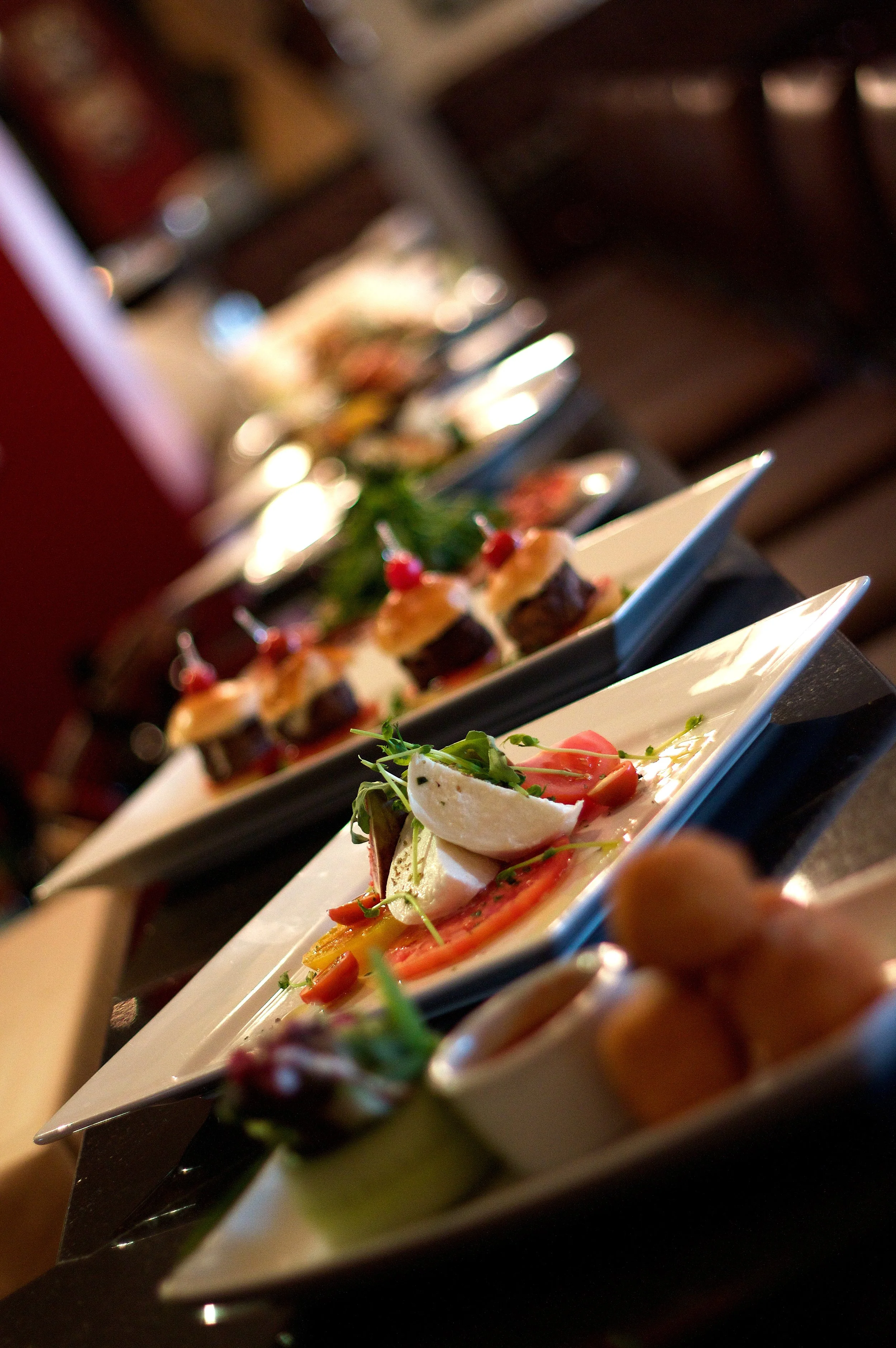 Assorted appetizers on white rectangular plates, including a Caprese salad with tomato, mozzarella, and basil, and small savory canapés with toppings, served on a dark table in a restaurant setting.