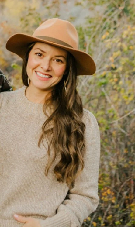 A woman with long wavy brown hair wearing a beige sweater and a wide-brimmed tan hat, smiling outdoors with fall foliage in the background.