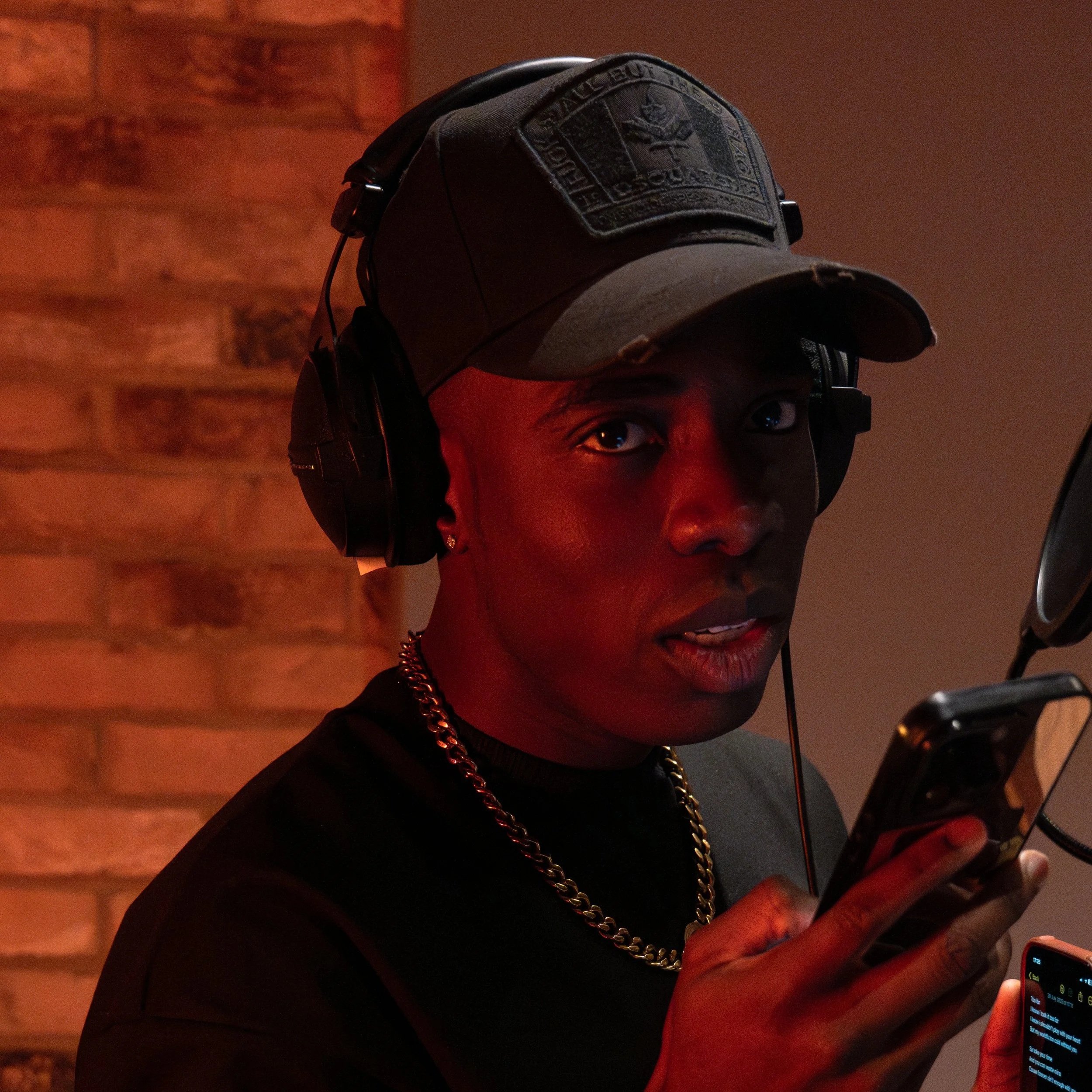 A young person wearing a baseball cap, headphones, and a gold chain, holding a smartphone, standing against a brick wall background.