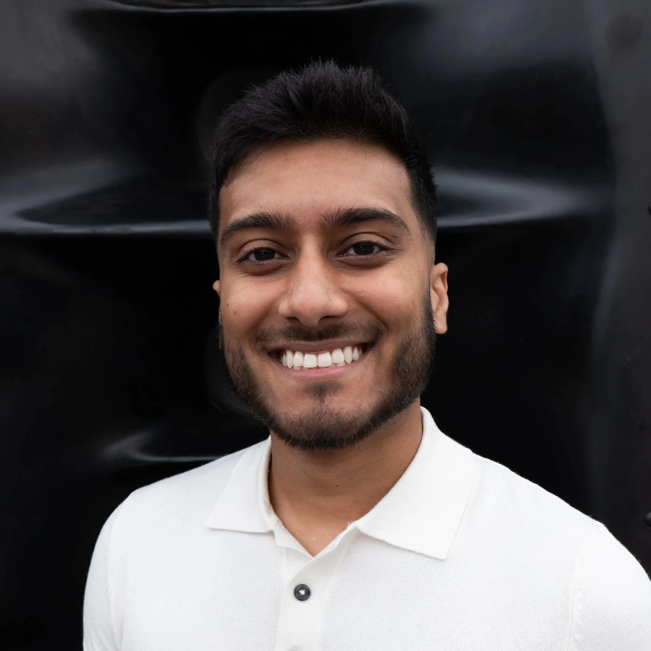 A young man with dark hair and a beard smiling at the camera, wearing a white polo shirt, against a black background.