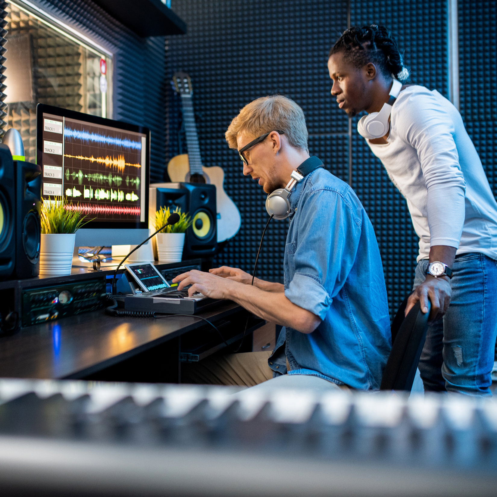 Two men in a music studio working on audio mixing. One seated at a computer with headphones around his neck and working on a digital audio workstation. The other standing beside him, observing. The studio has soundproof walls, musical instruments, and audio equipment.