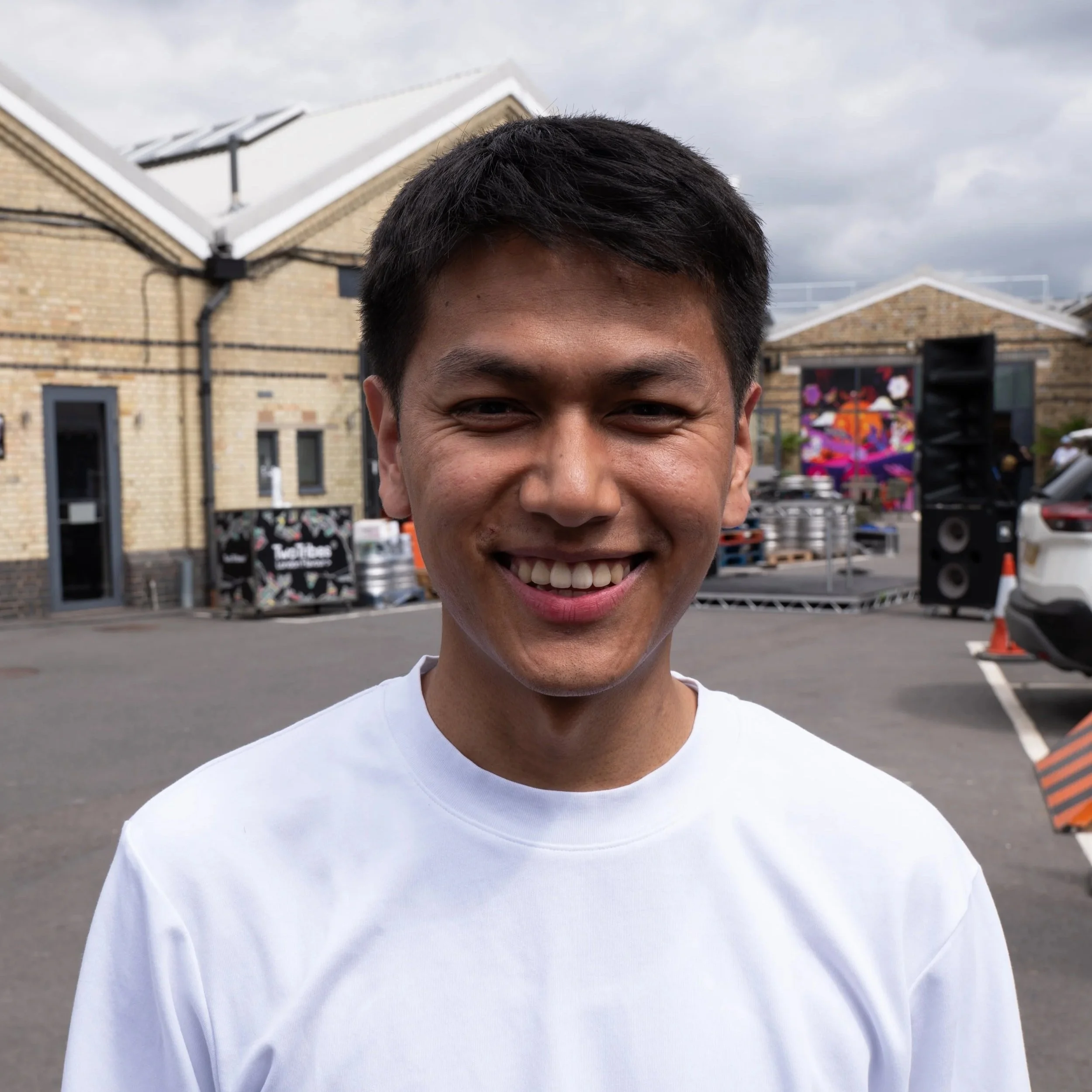 Young man smiling outdoors at event with buildings, stage, and equipment in the background