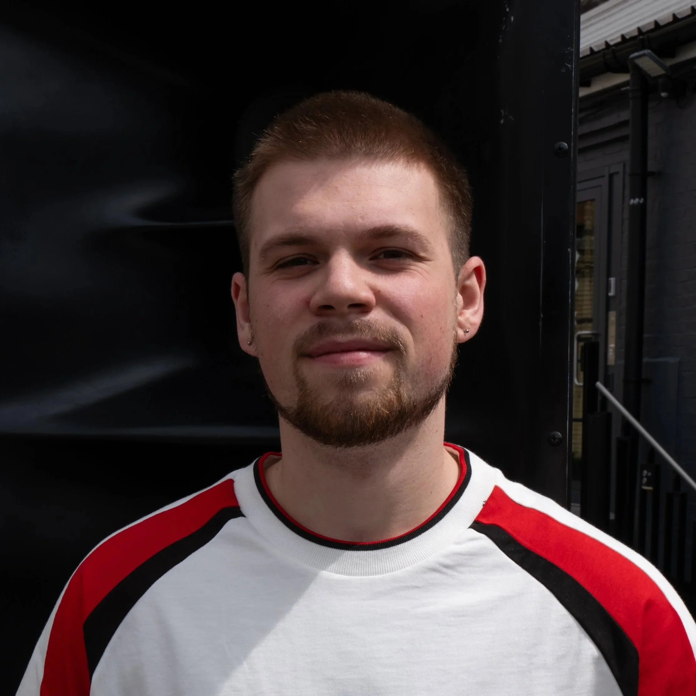 A young man with short brown hair, facial hair, and earrings wearing a white, red, and black shirt standing outdoors with a dark background behind him.