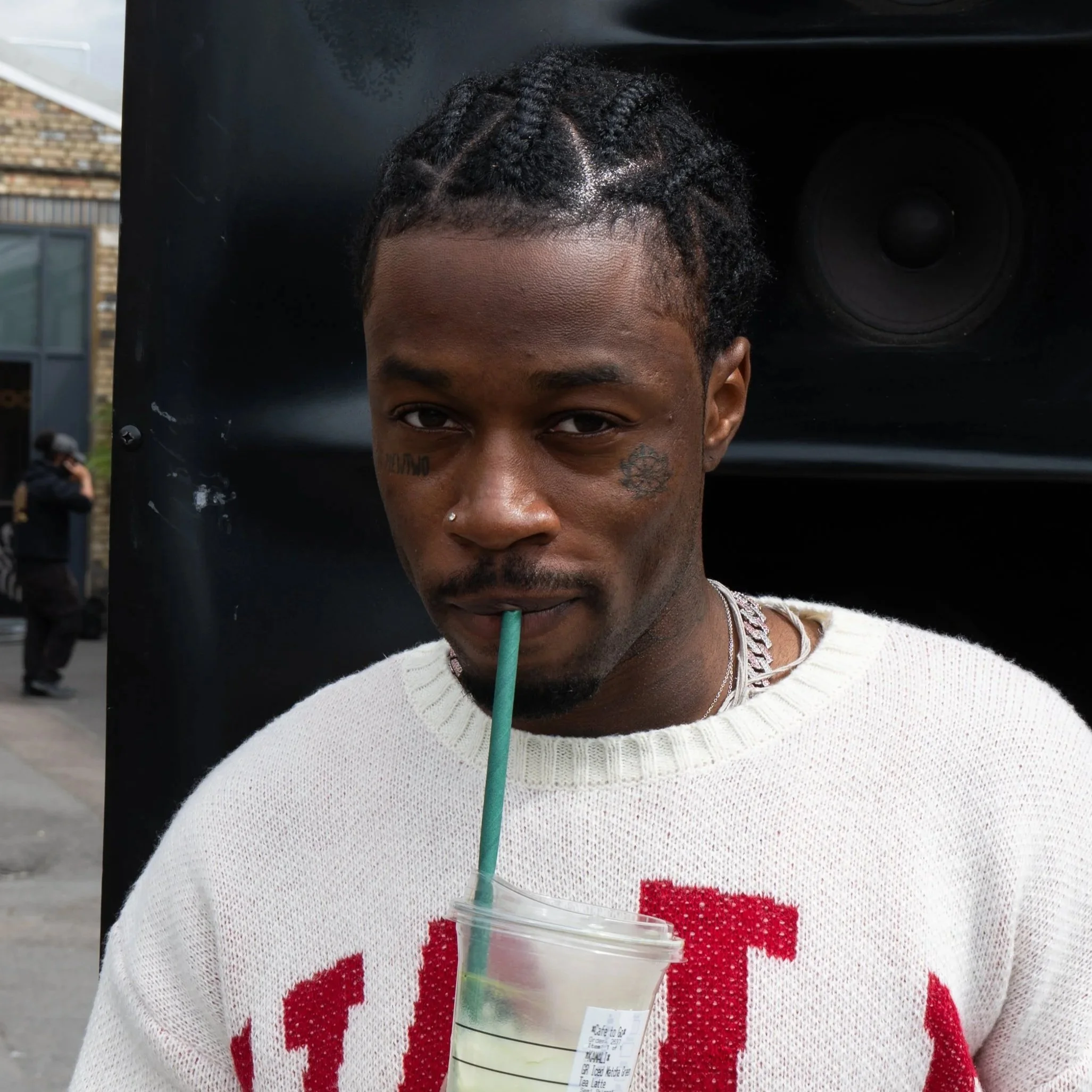 A young man with braided hair drinking from a cup using a straw, wearing a white sweatshirt with red lettering, standing outdoors near a large black speaker.