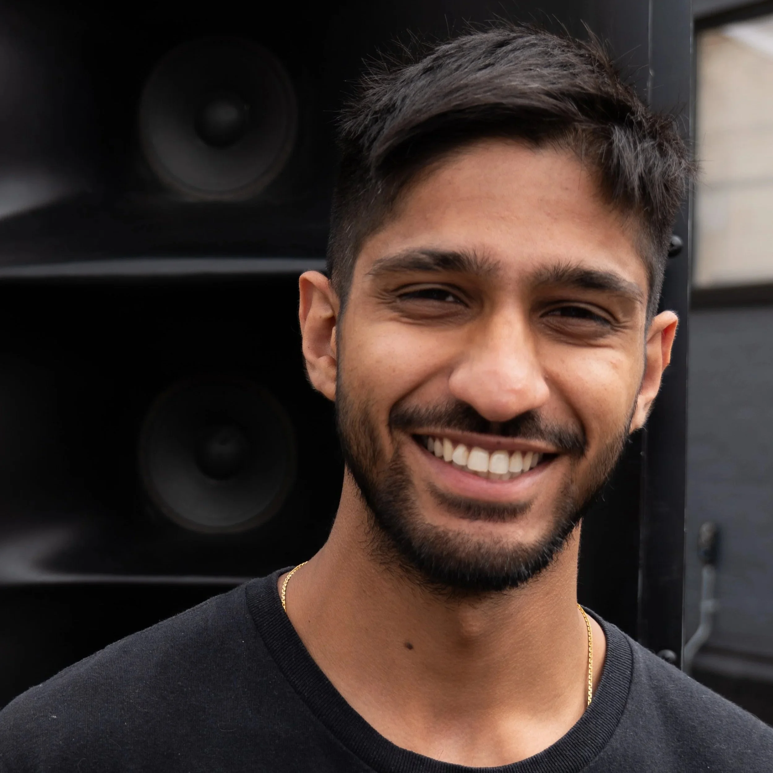 A smiling man with dark hair and a beard standing outdoors in front of large black speakers.