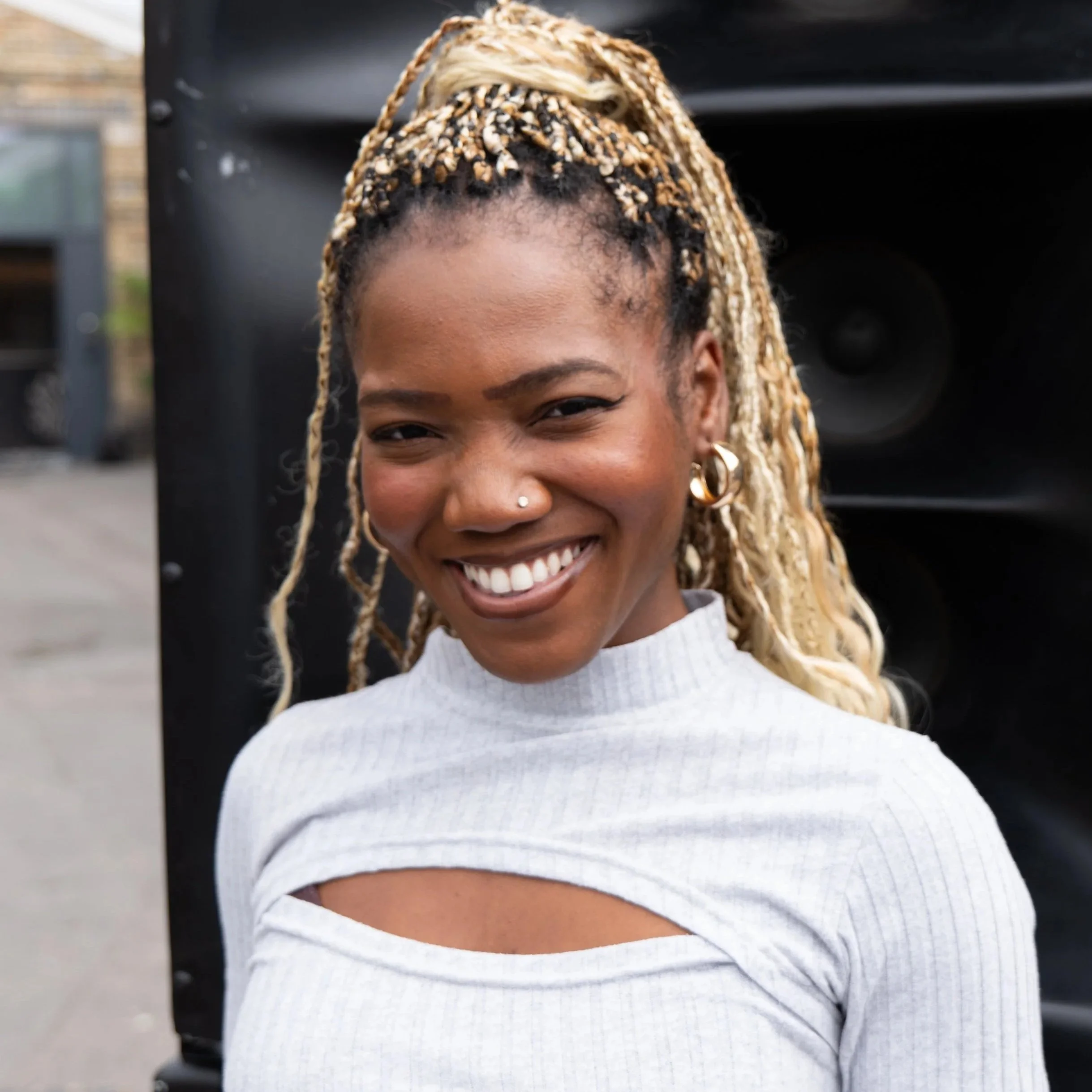 A smiling woman with blonde braided hair, wearing hoop earrings and a light-colored top with a cutout design.