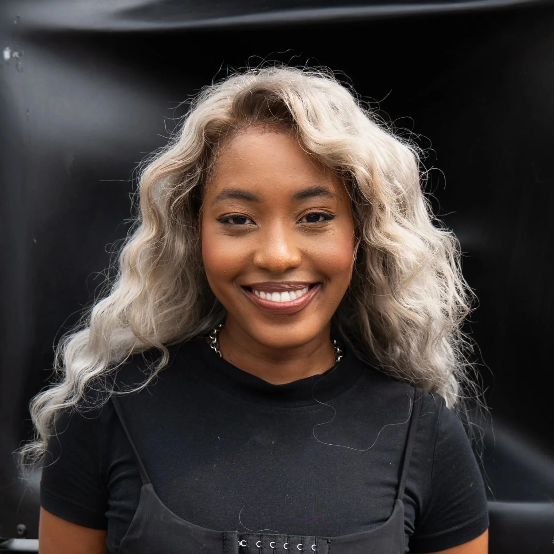A smiling young woman with long, curly, blonde hair, wearing a black top and a black chest strap, standing against a black background.