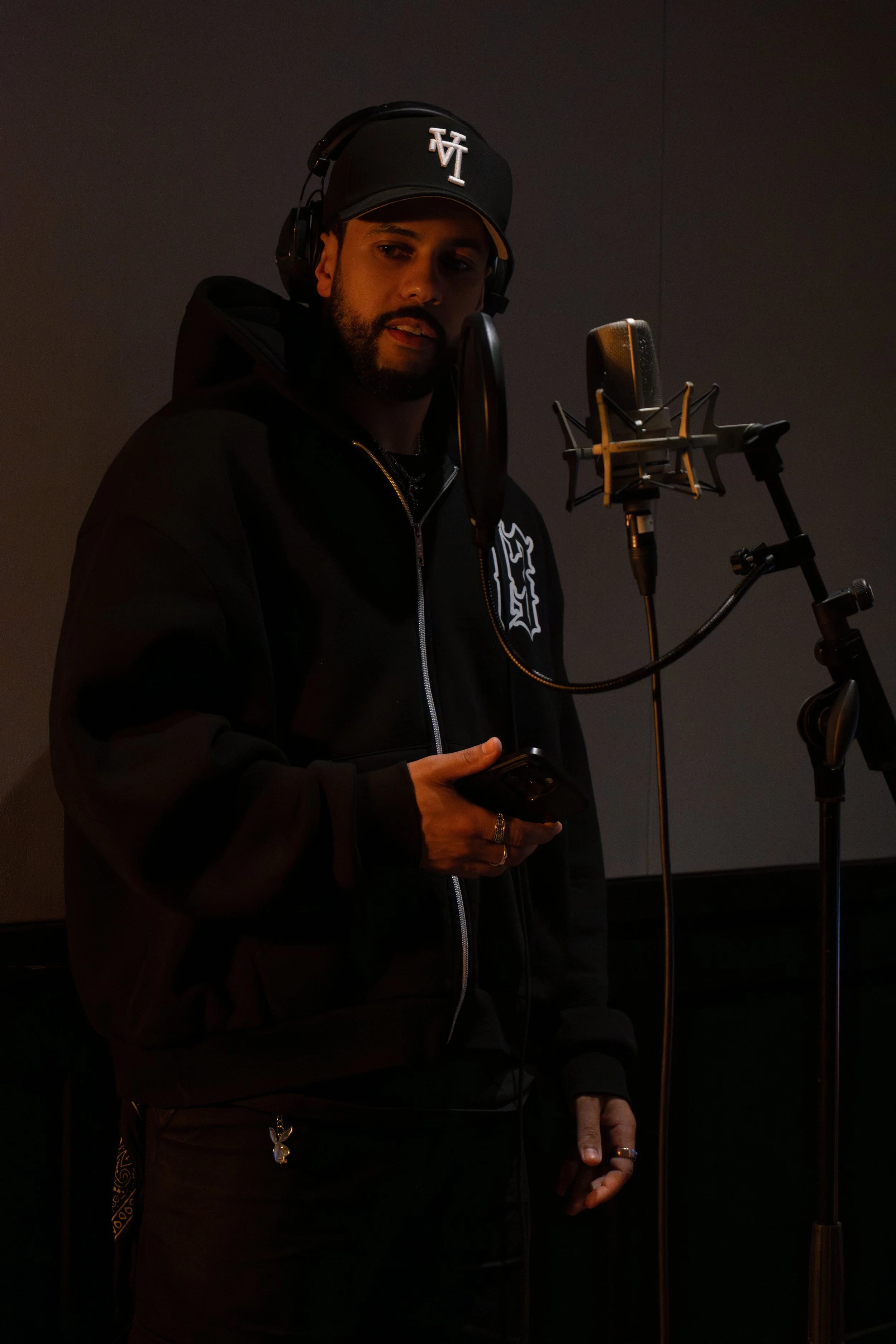 A man in a black hoodie and black cap with white logo stands in a recording studio, holding a smartphone, and positioned at a microphone with a pop filter, headphones on his head.