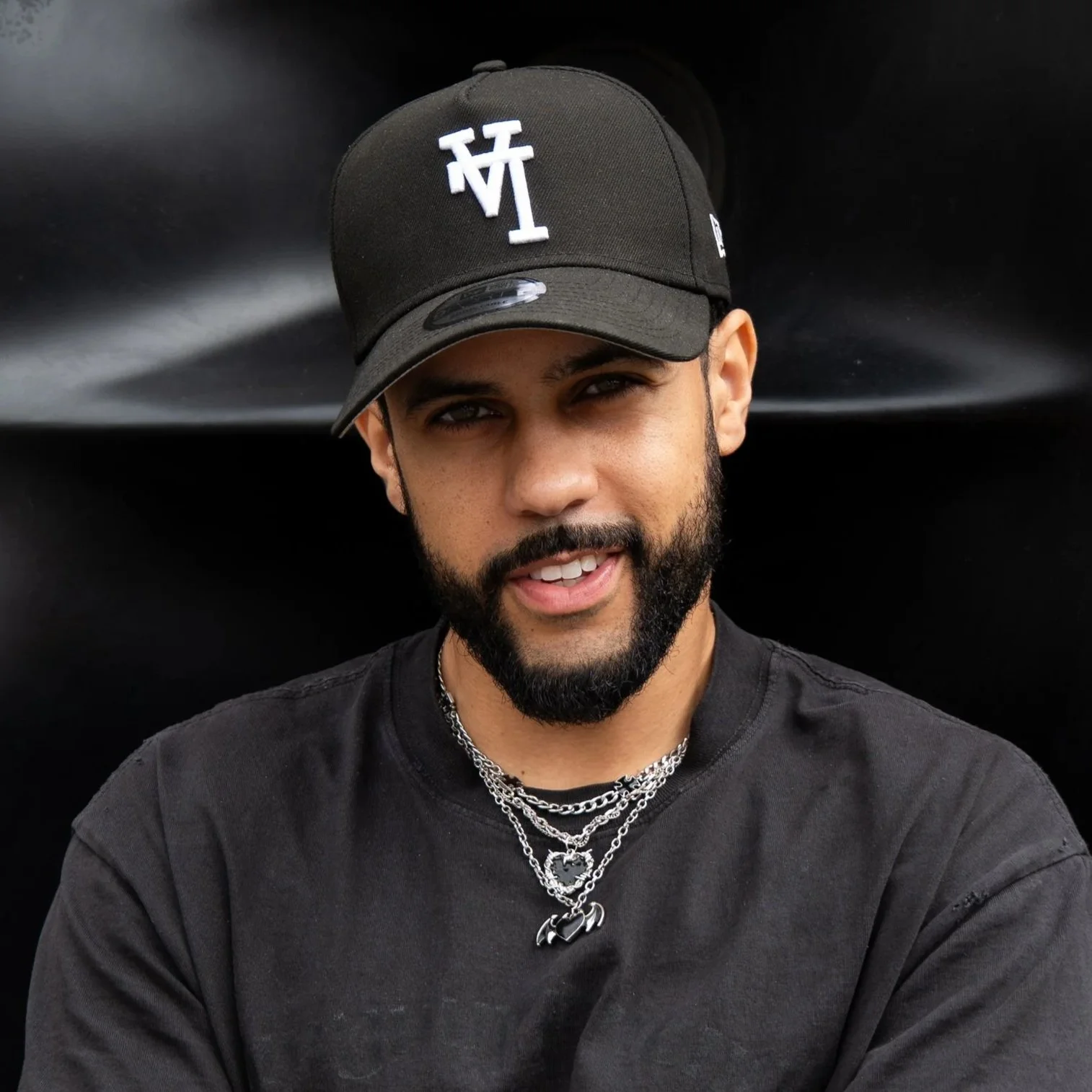 A young man with a beard, wearing a black baseball cap and black shirt, posing against a black background.