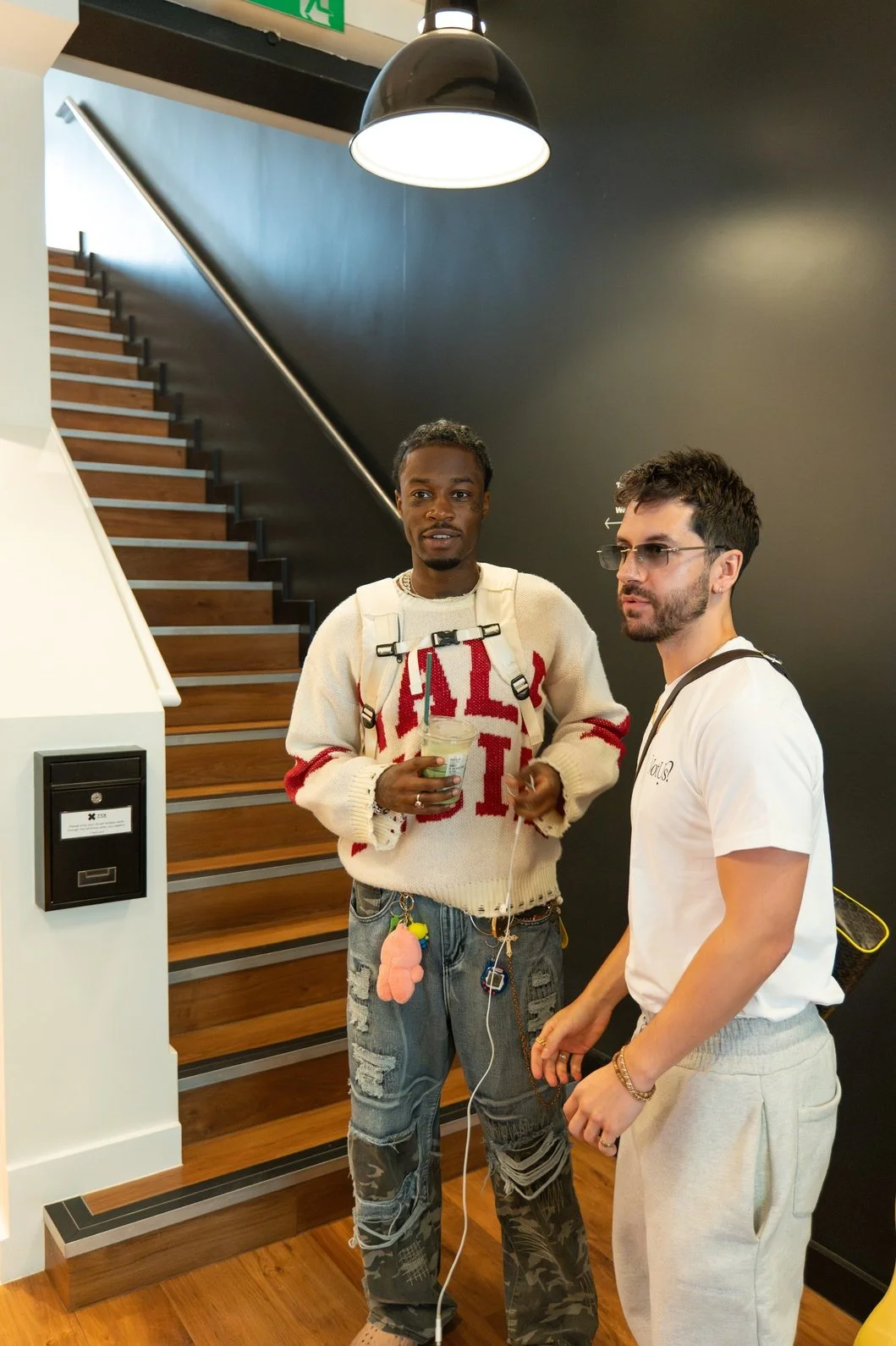 Two men standing near a staircase, engaged in conversation. One of them is holding a drink, wearing a Kellogg's sweater and distressed jeans; the other is dressed in a white t-shirt and light-colored pants, with glasses and a wrist bracelet.